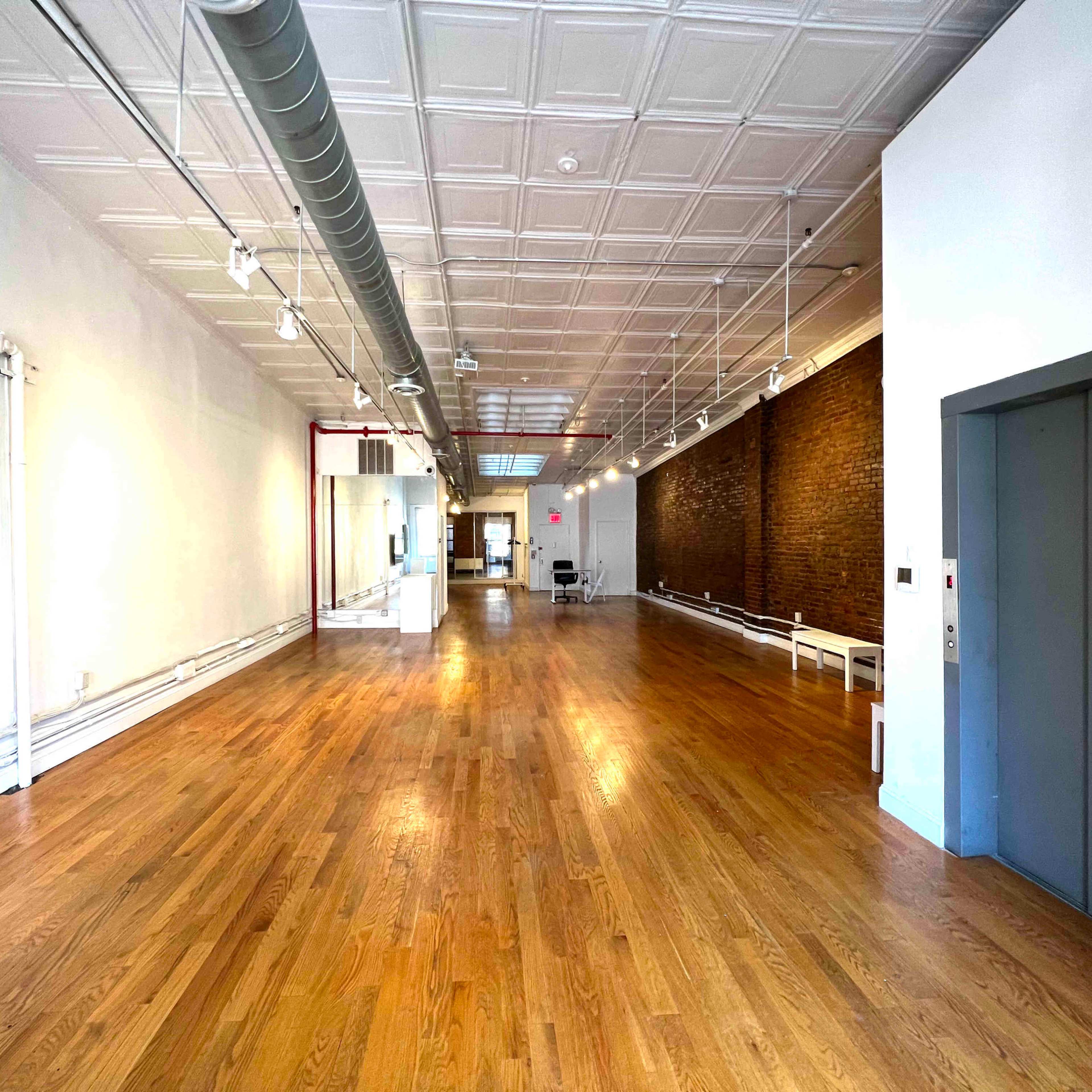 A spacious, well-lit interior with wooden flooring, exposed brick walls, and an elevator on the right side.