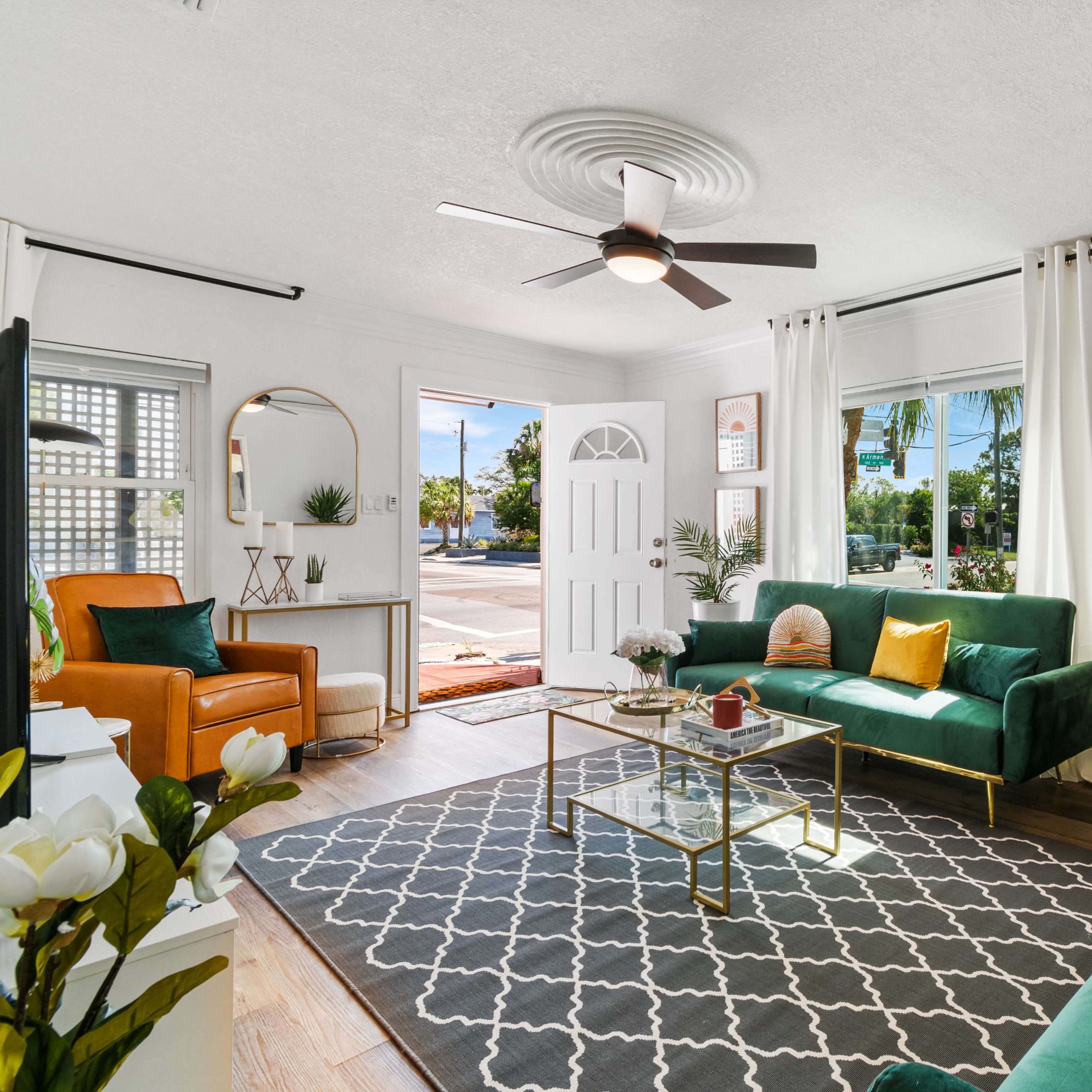 A bright living room with green sofas, a coffee table, a ceiling fan, and large windows providing natural light.