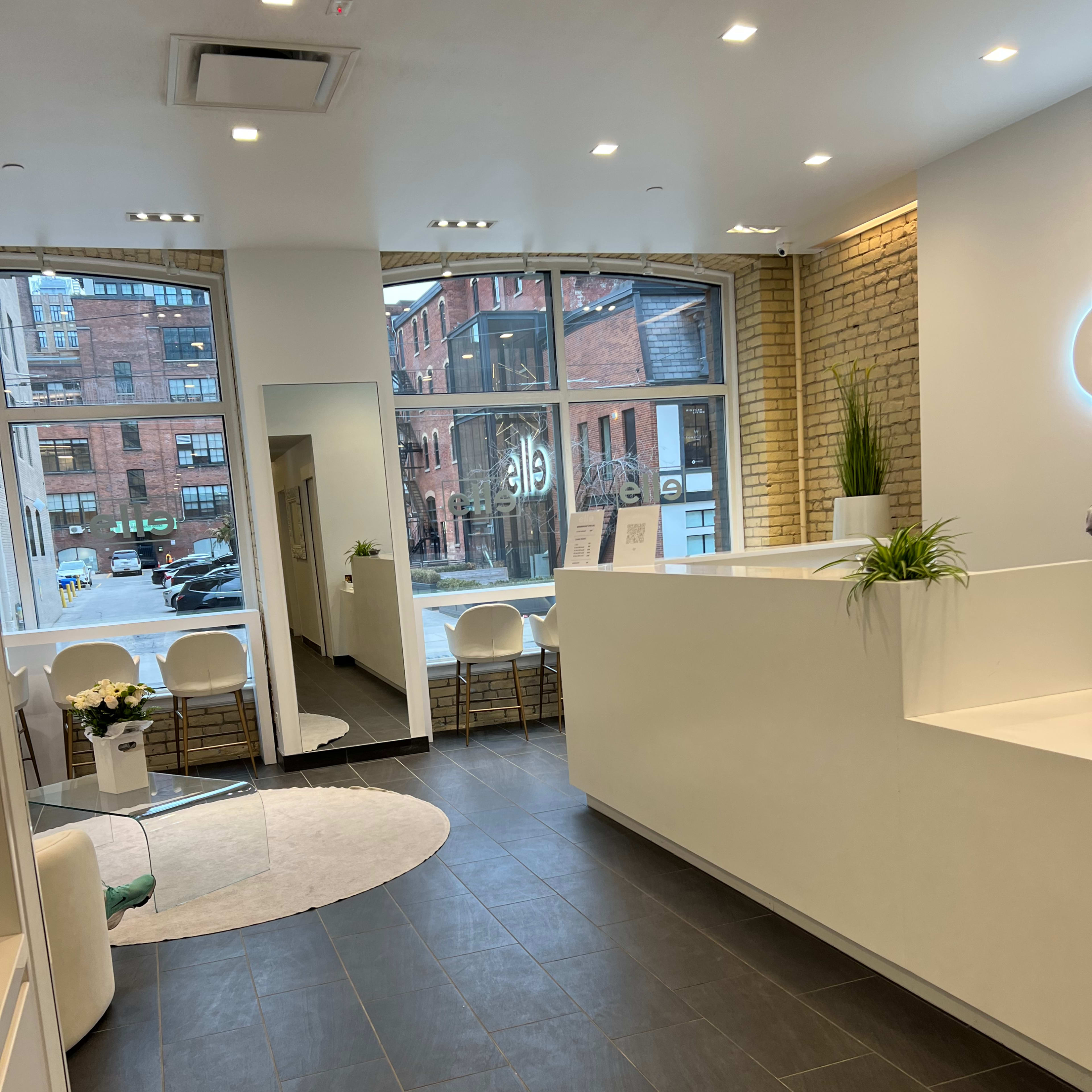 A modern, well-lit reception area with a light-colored wall, a circular rug, and several chairs, along with large windows revealing a view of a city street.