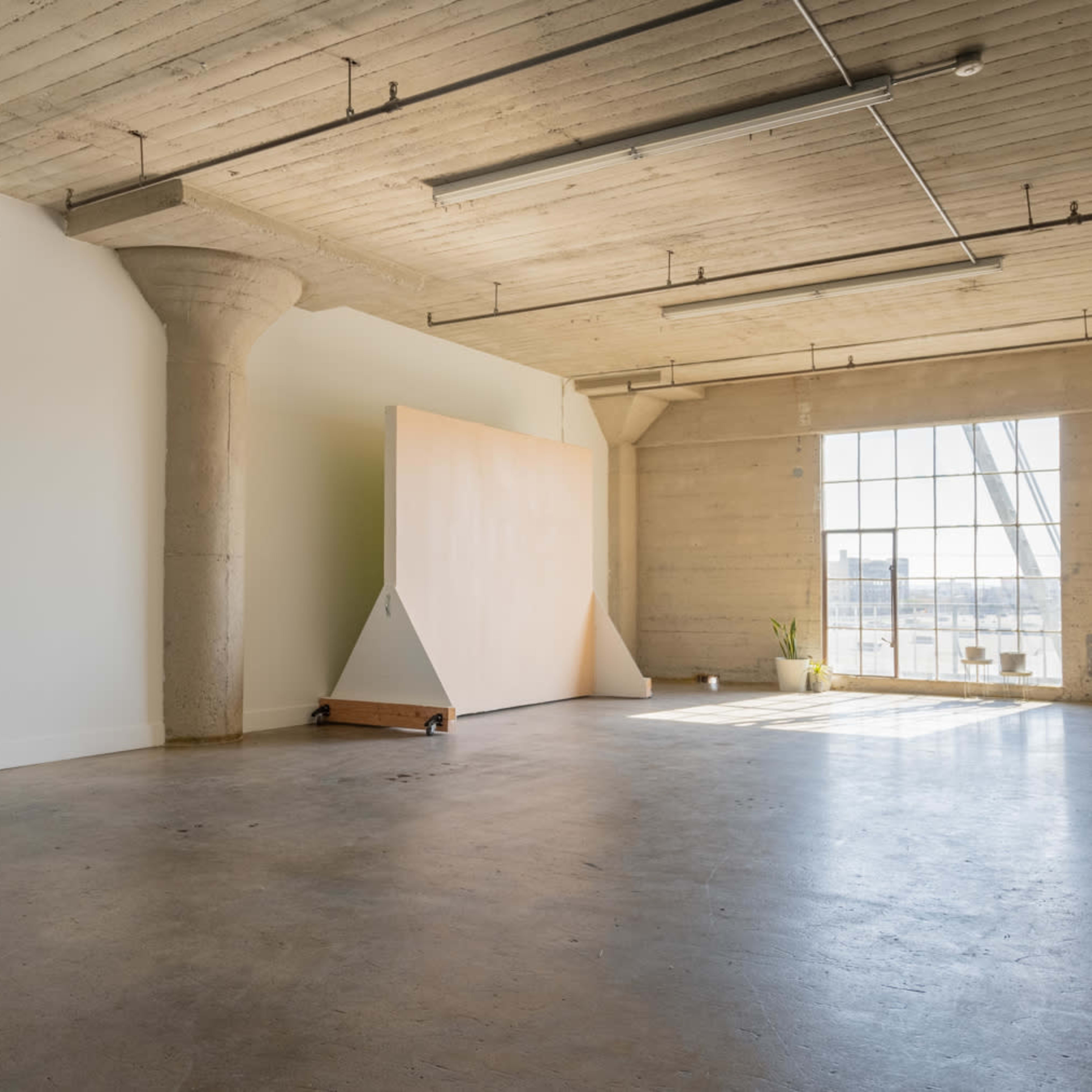 A spacious, empty room with concrete walls, high ceilings, and a large window letting in natural light, featuring a large, triangular wooden panel against one wall.