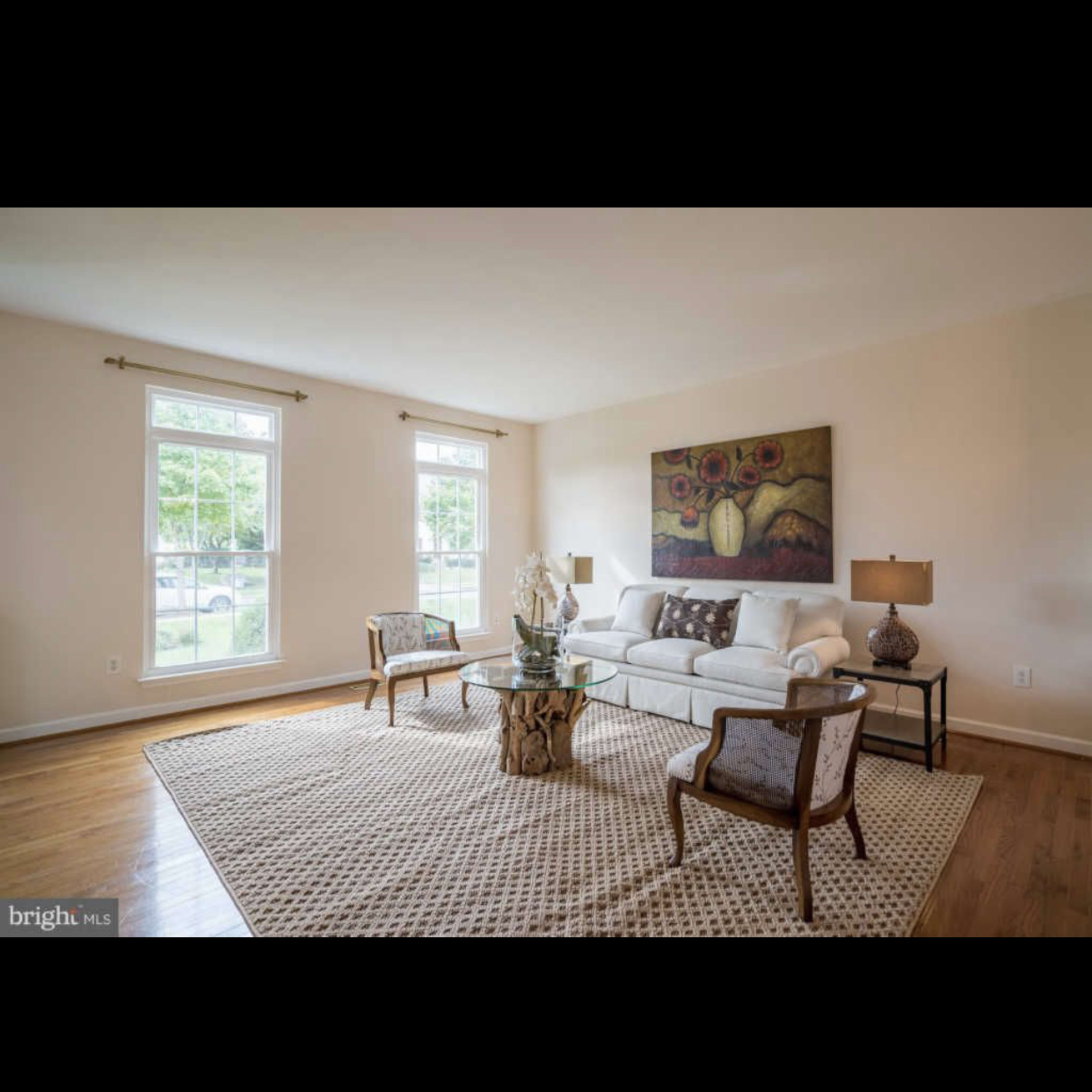 A minimalist living room featuring a white sofa, two wooden chairs, a large area rug, and a central coffee table with a plant, illuminated by natural light from two windows.