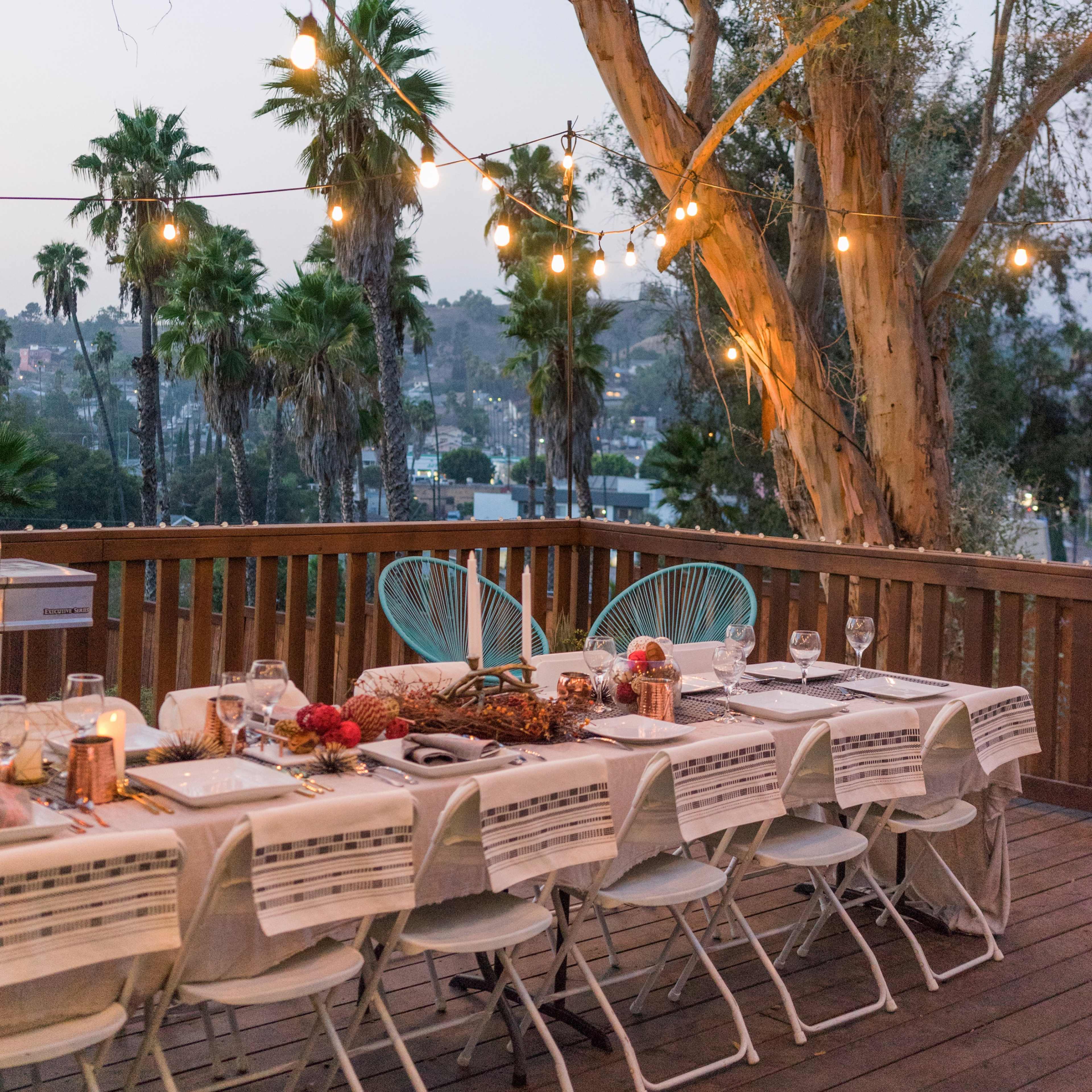 A long dining table is set on a deck under string lights, surrounded by palm trees and overlooking a scenic view at dusk.
