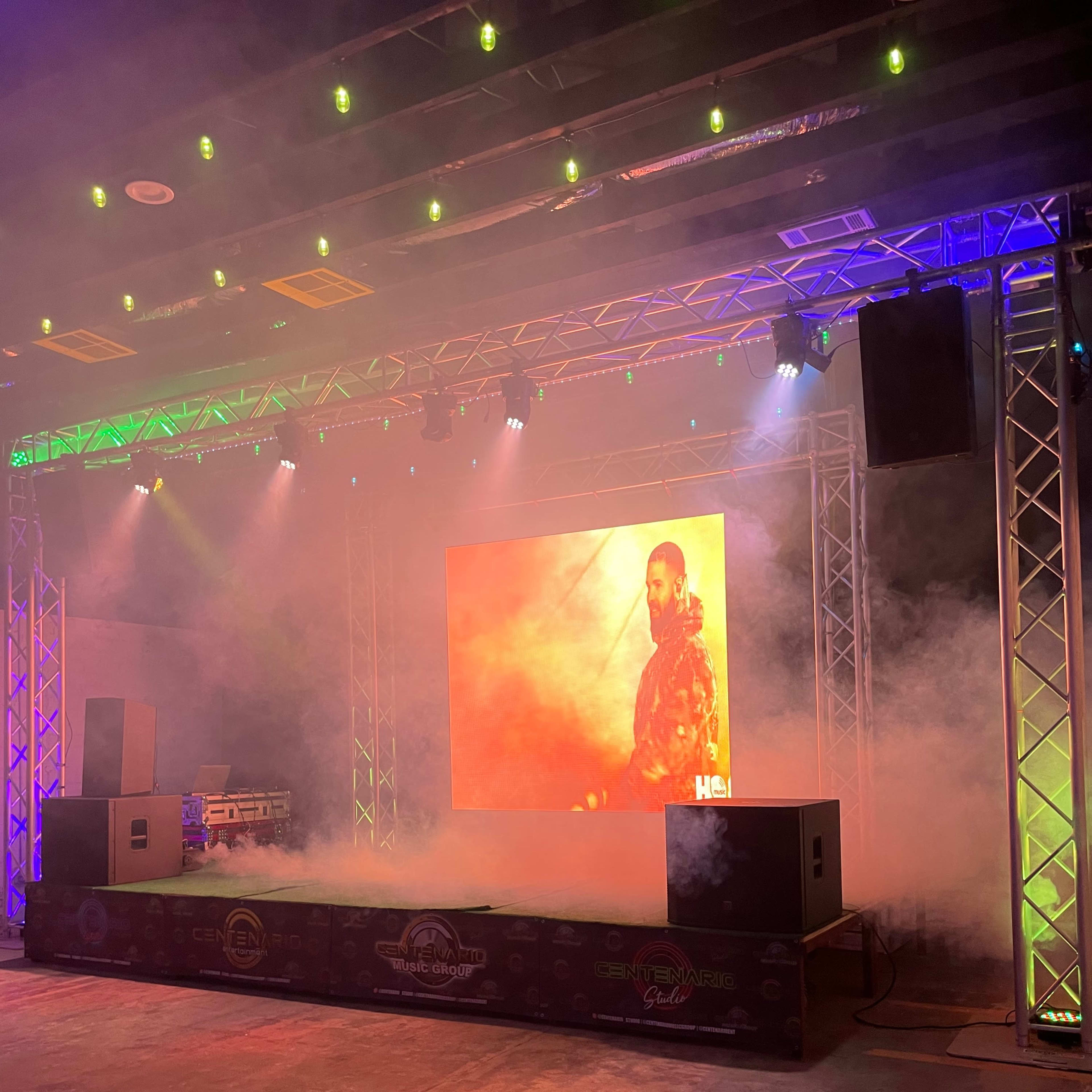 A stage setup features illuminated trussing, smoke effects, and a large backdrop displaying a performer.