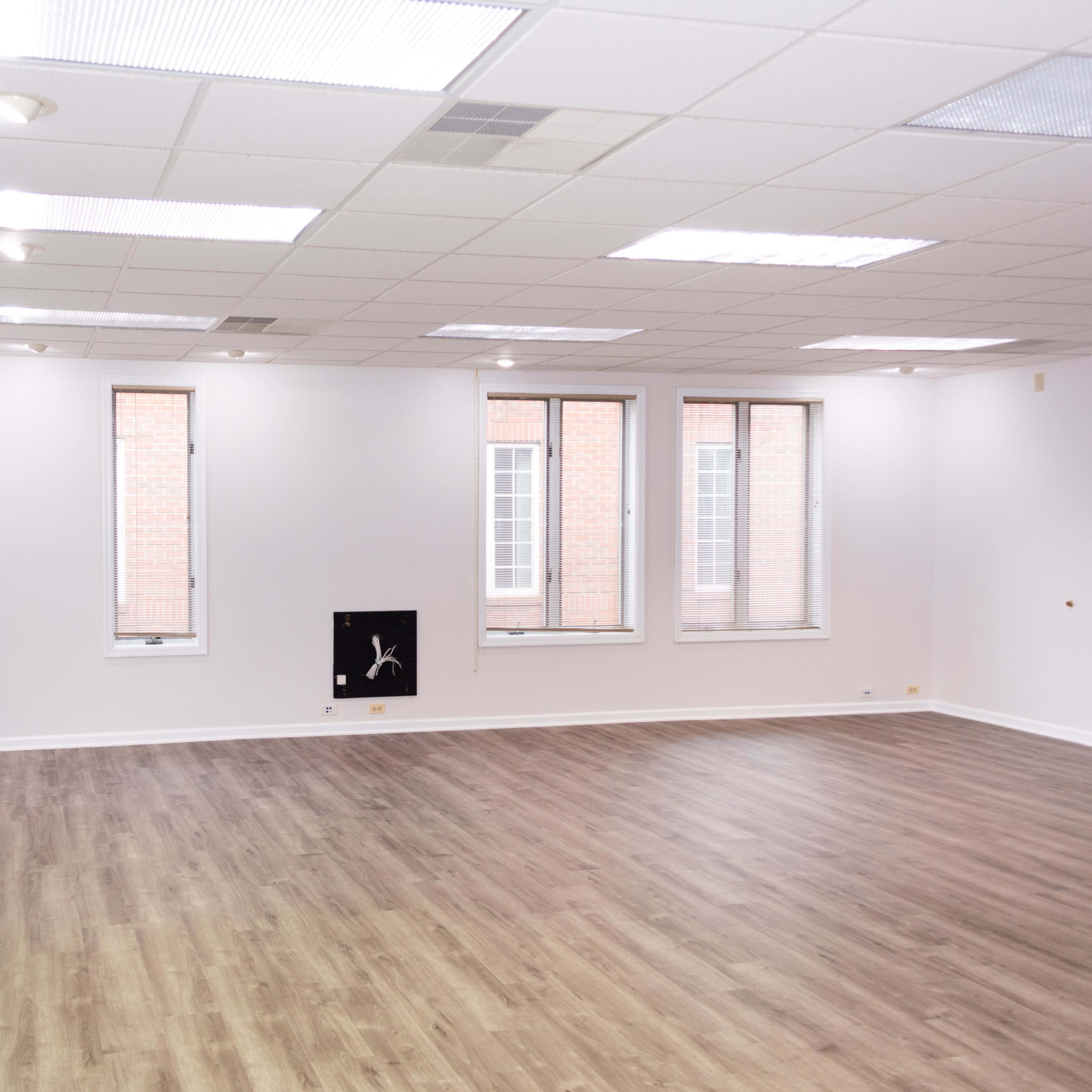An empty room with wooden flooring and three windows, featuring plain white walls and a small wall-mounted object.