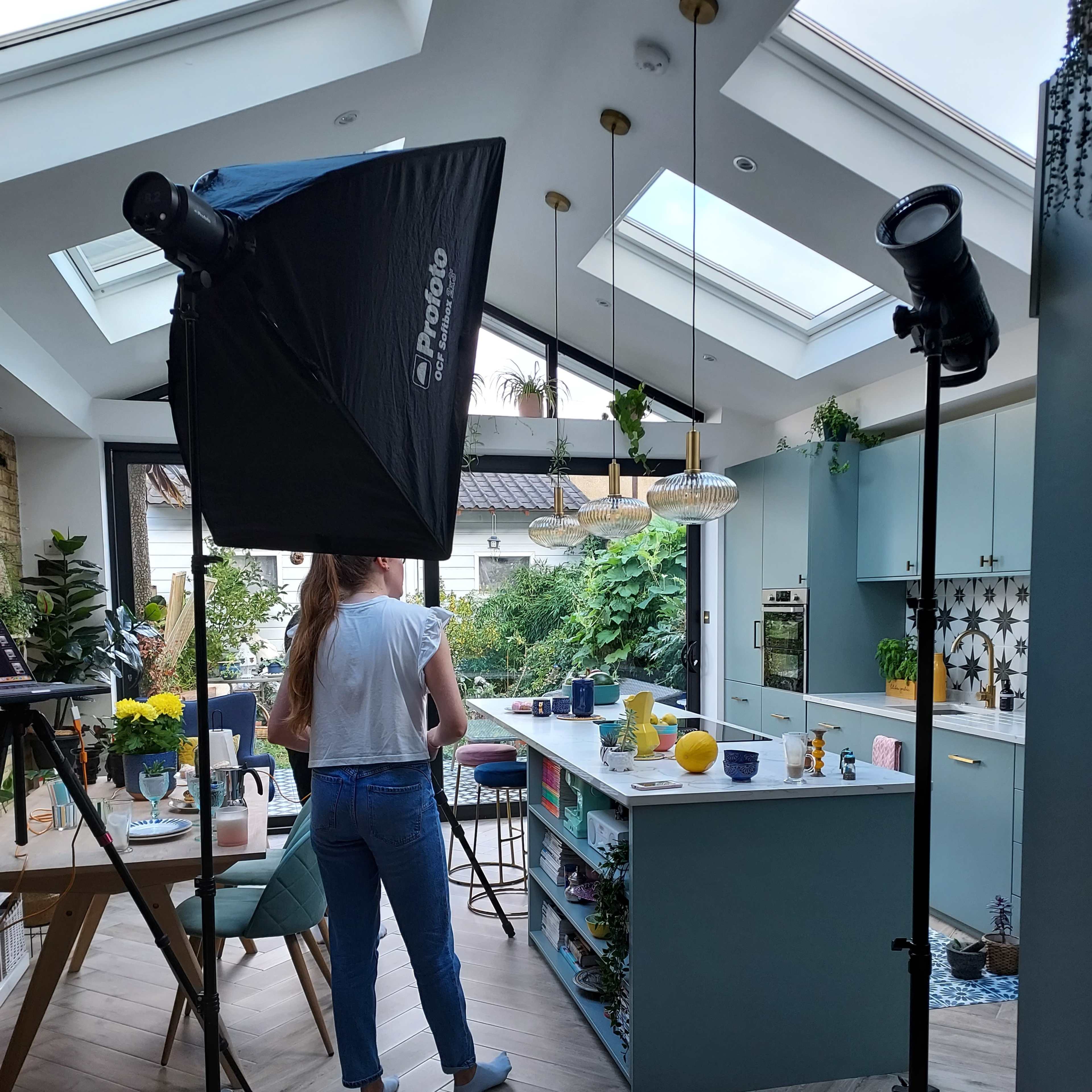 A person stands near a kitchen island in a modern kitchen, illuminated by softbox lighting and surrounded by various kitchen items and plants.