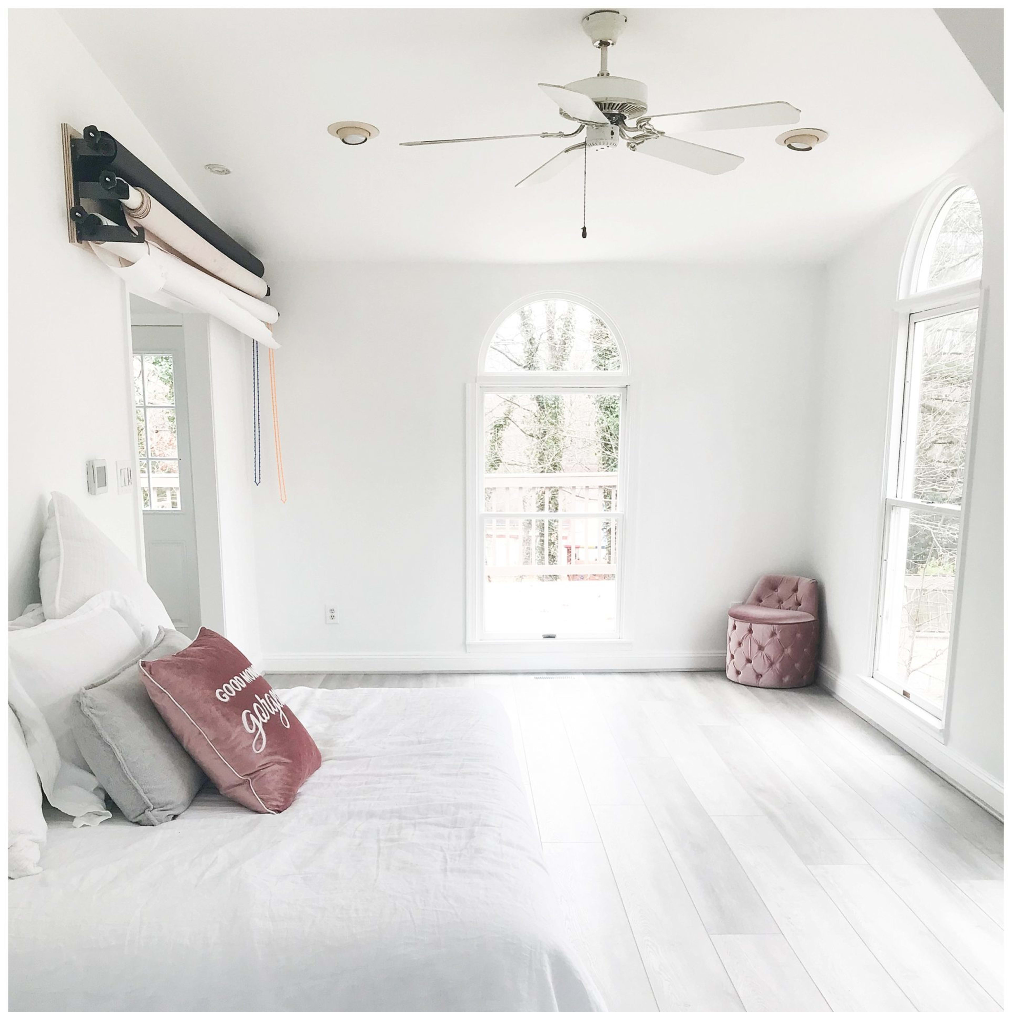 A well-lit room features a white bed with a decorative pillow, a pink upholstered chair, and large windows overlooking a balcony.