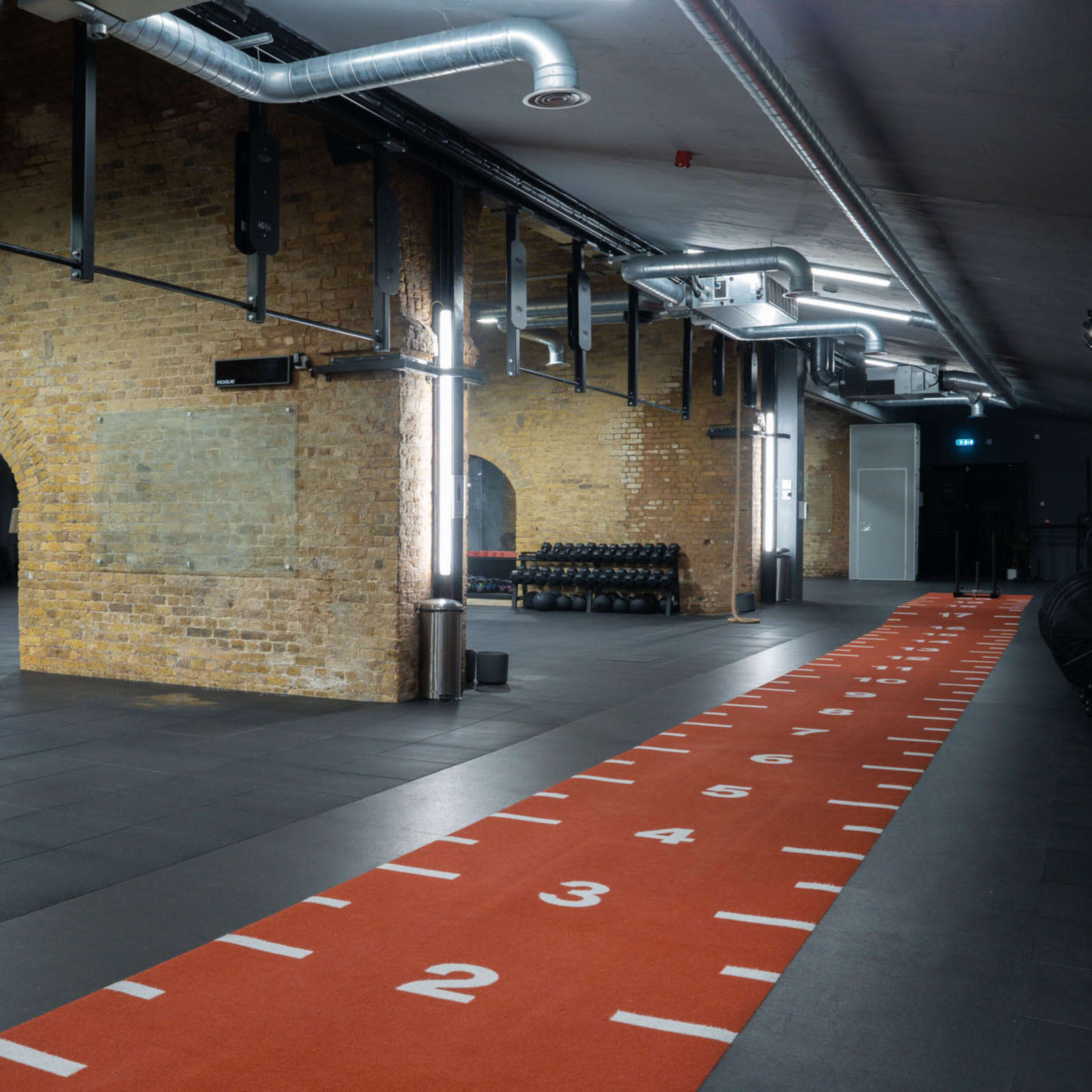 A spacious gym interior featuring exposed brick walls, a gray tiled floor, and an orange track marked with numbers on one side.