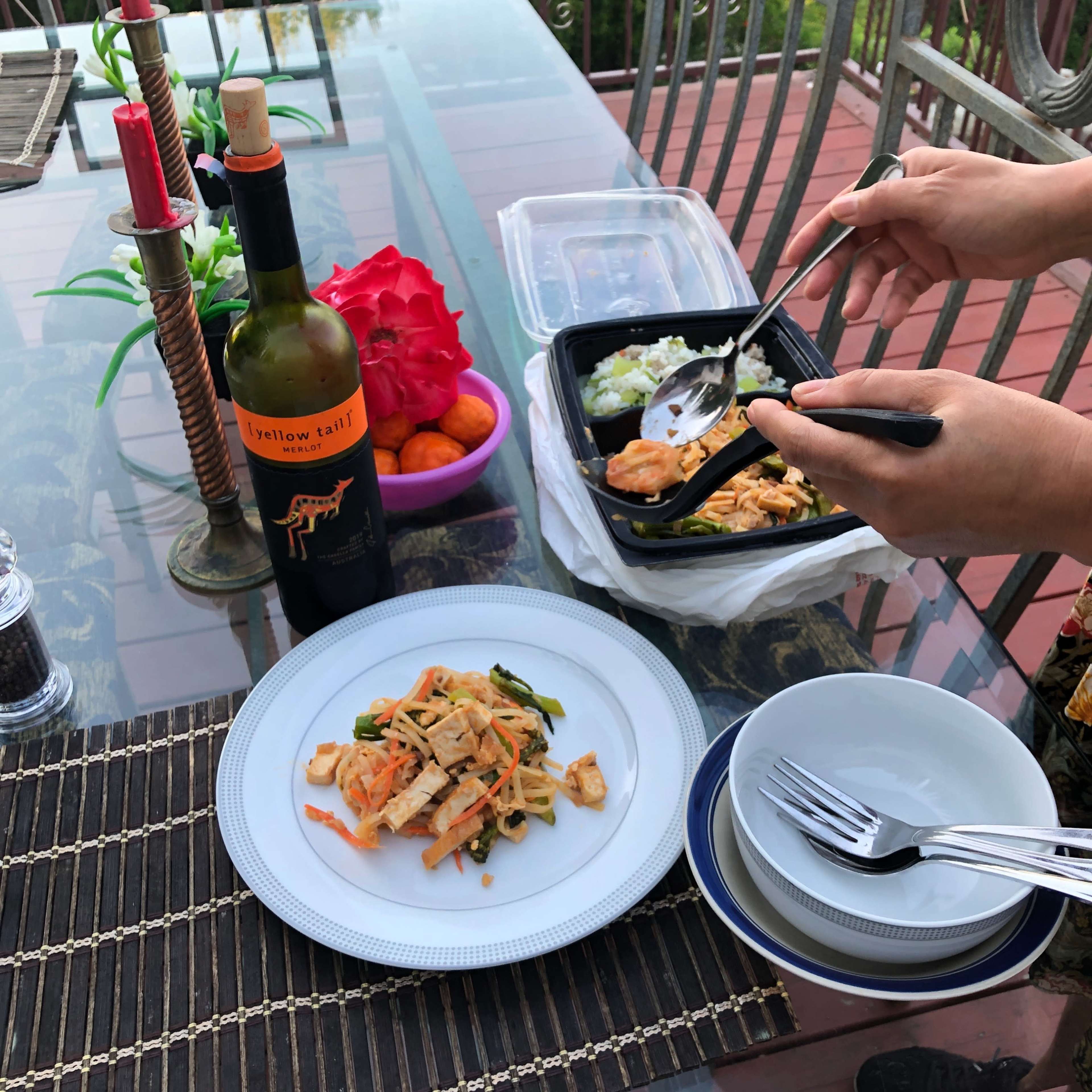 A woman serves food from a takeout container onto a plate while a bottle of wine and decorative items are arranged on a glass table.