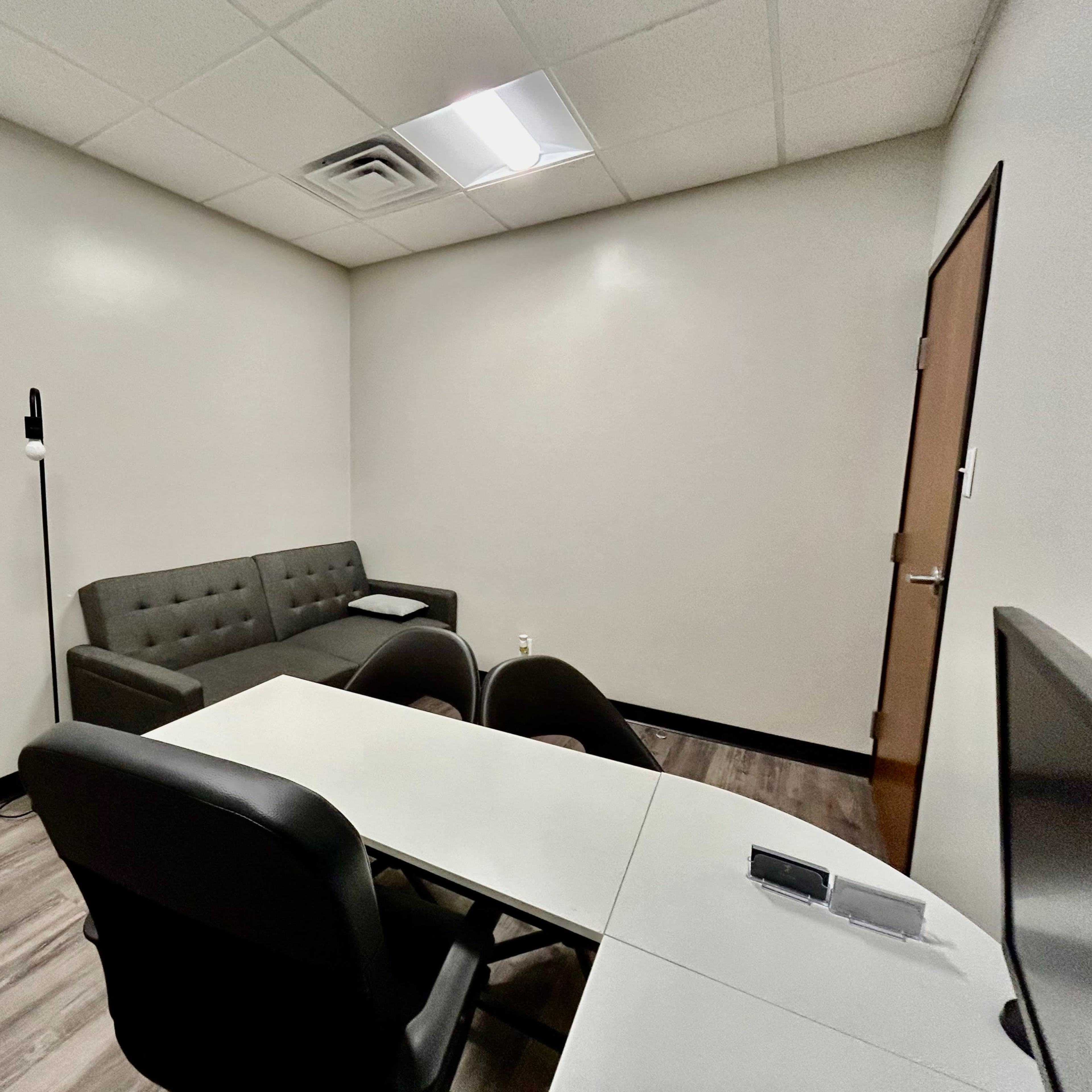 A small office or meeting room featuring a grey couch, a table with chairs, and a door.