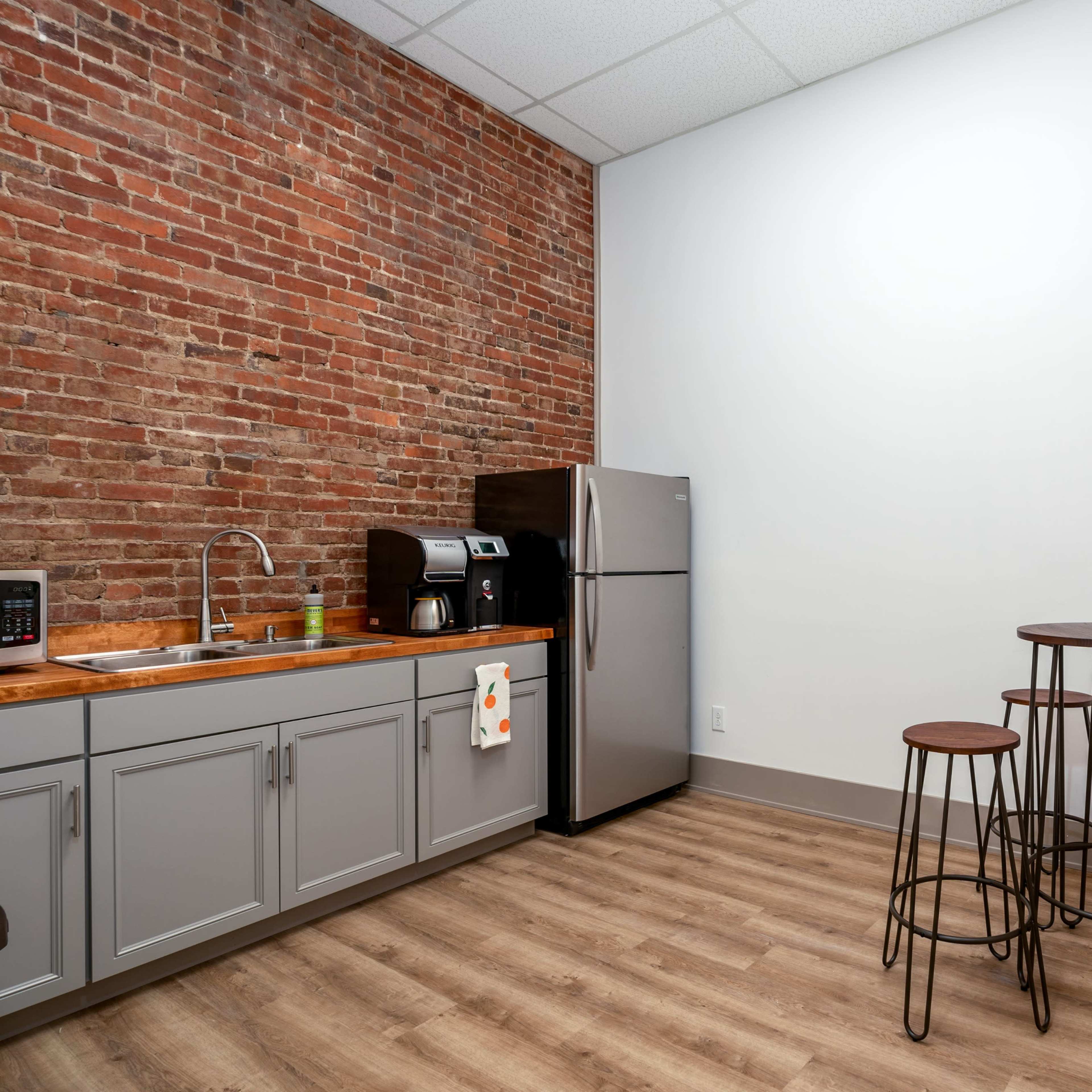 A minimalist kitchen area with exposed brick walls, a stainless steel refrigerator, a wooden countertop, and a small dining set featuring three bar stools.