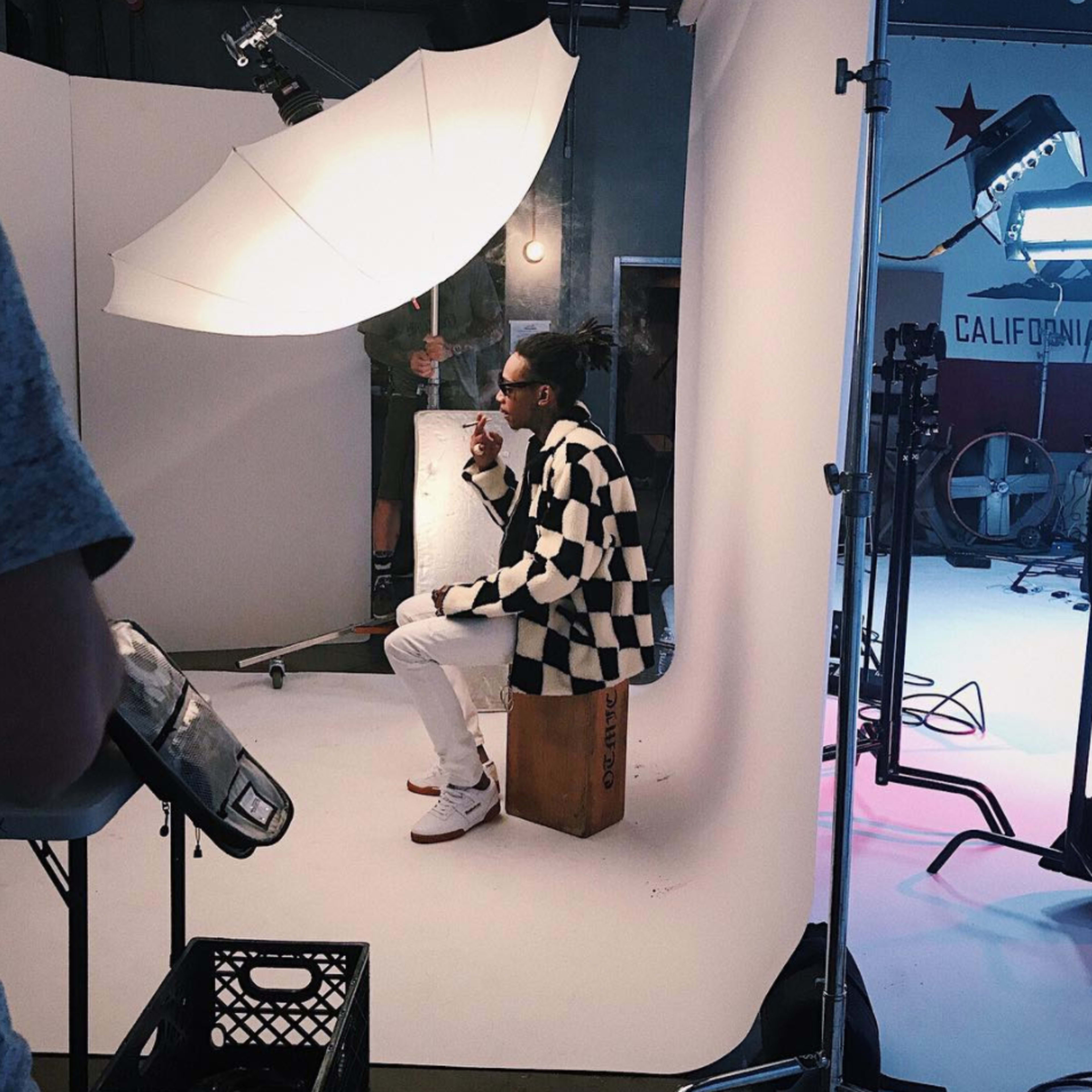 A model sits on a wooden crate in a photo studio, dressed in a checkered coat with a lighting umbrella positioned above.