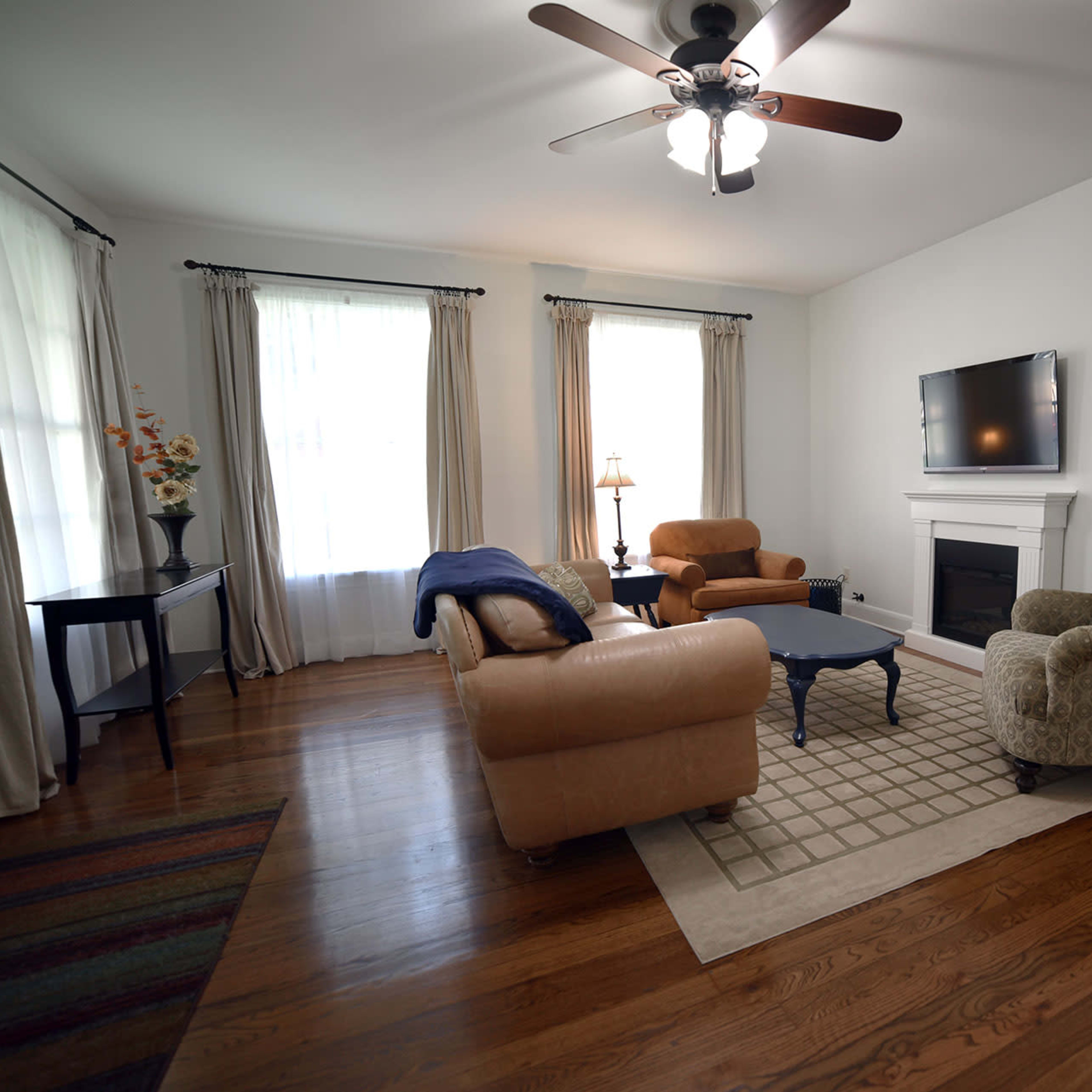 A living room with a ceiling fan, a sofa, two armchairs, a coffee table, a fireplace, and large windows dressed with curtains.