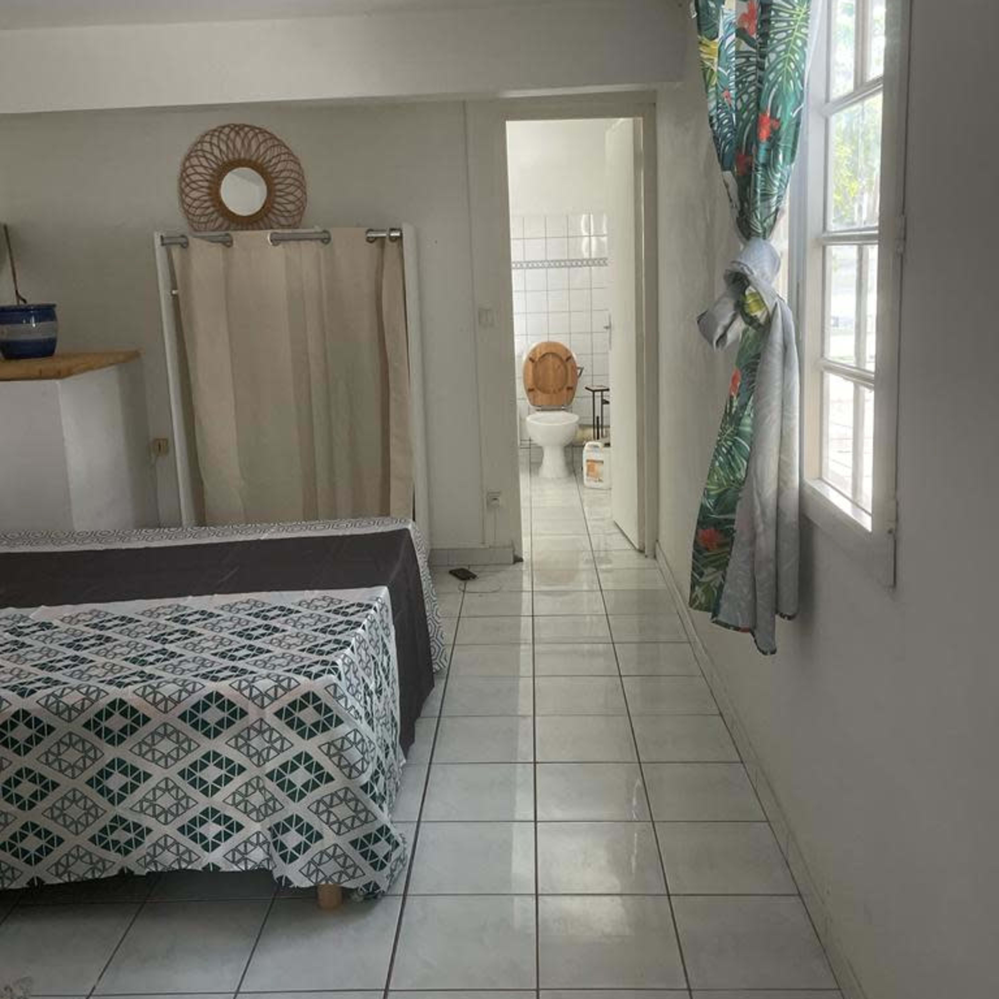 A simple room with a bed, a window draped with floral curtains, and a bathroom visible in the background.
