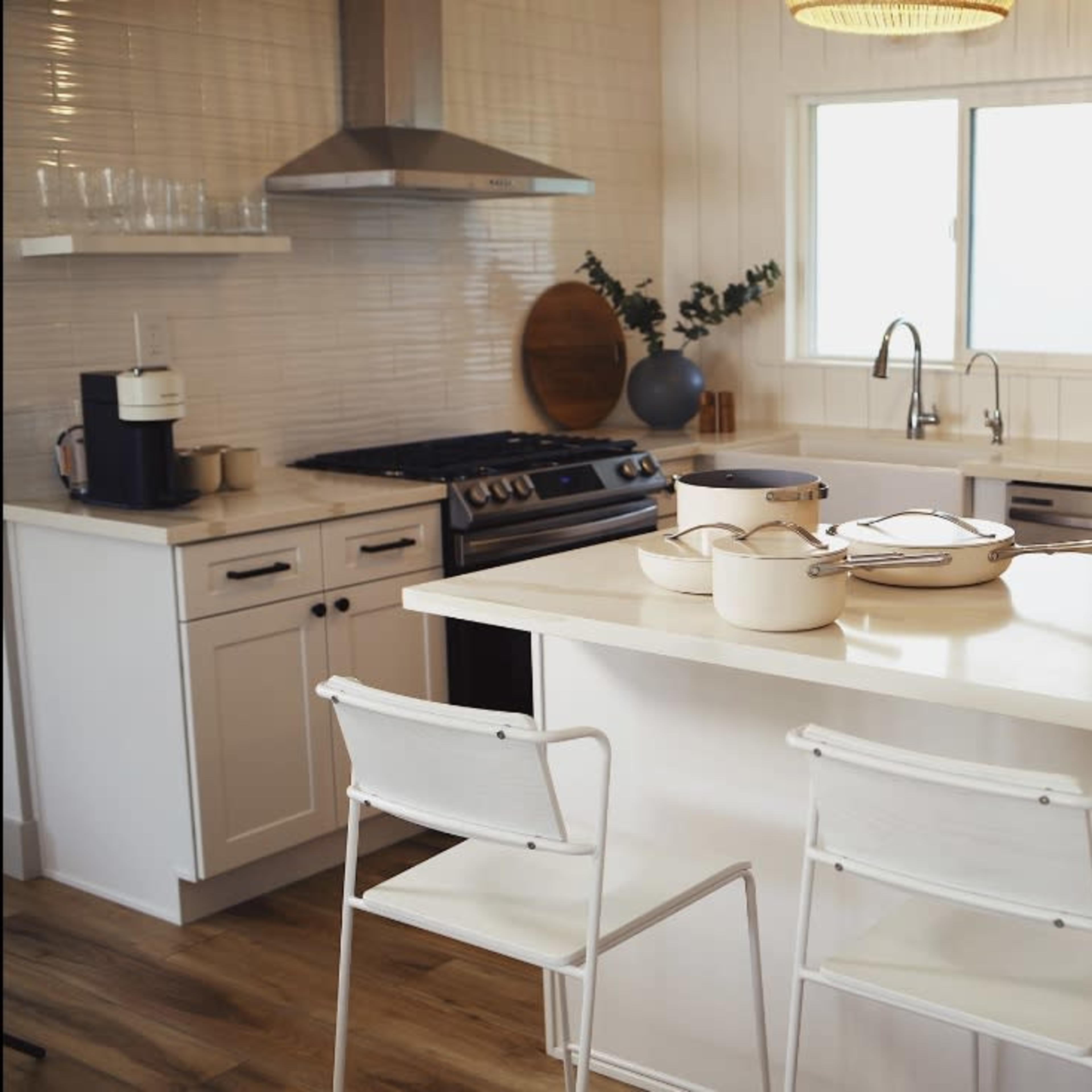 A modern kitchen features a stainless steel stove, a sink, and a small breakfast bar with two white chairs, topped with cooking pots and decorative plants.