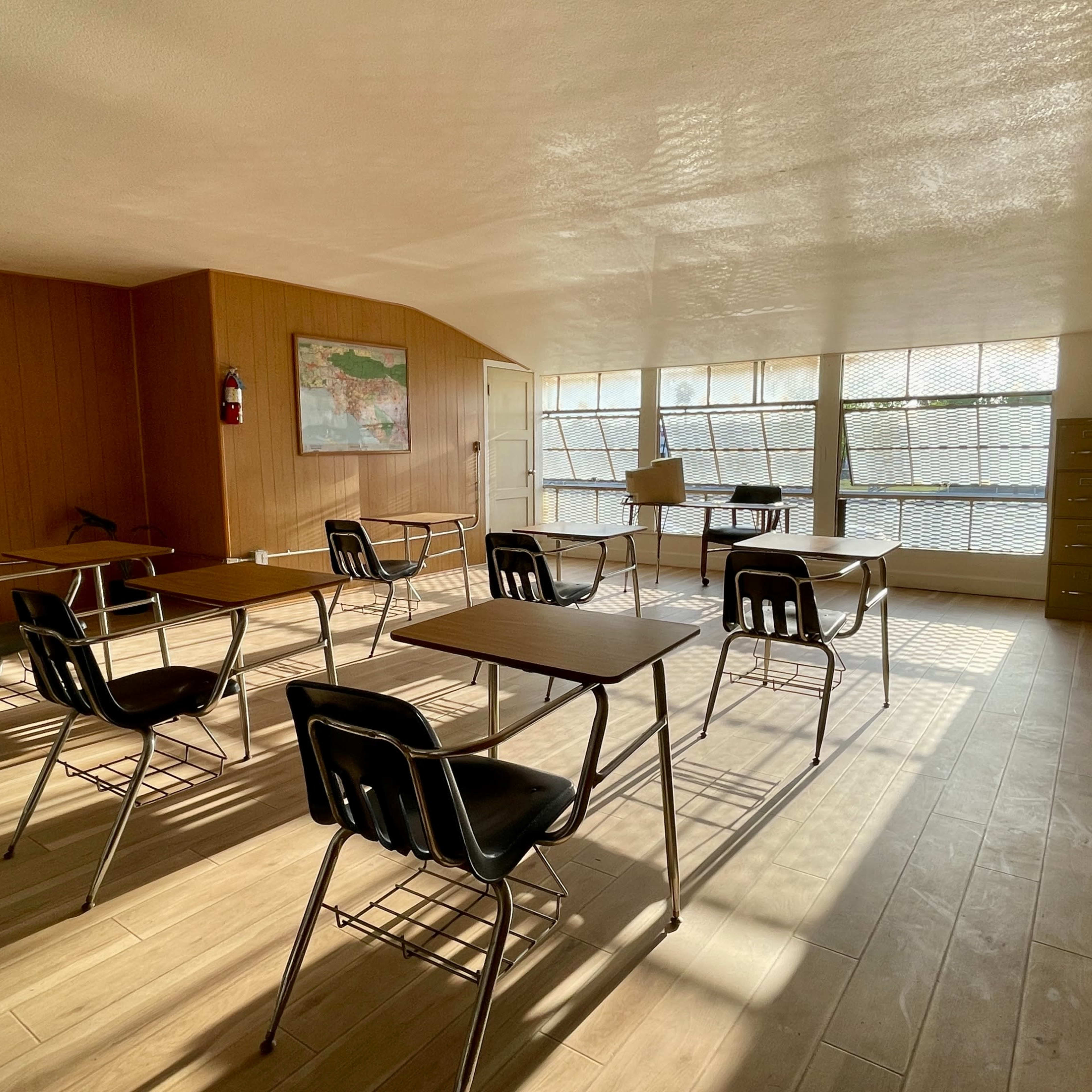A sparsely furnished classroom with several desks arranged in rows and a large window allowing natural light to fill the space.