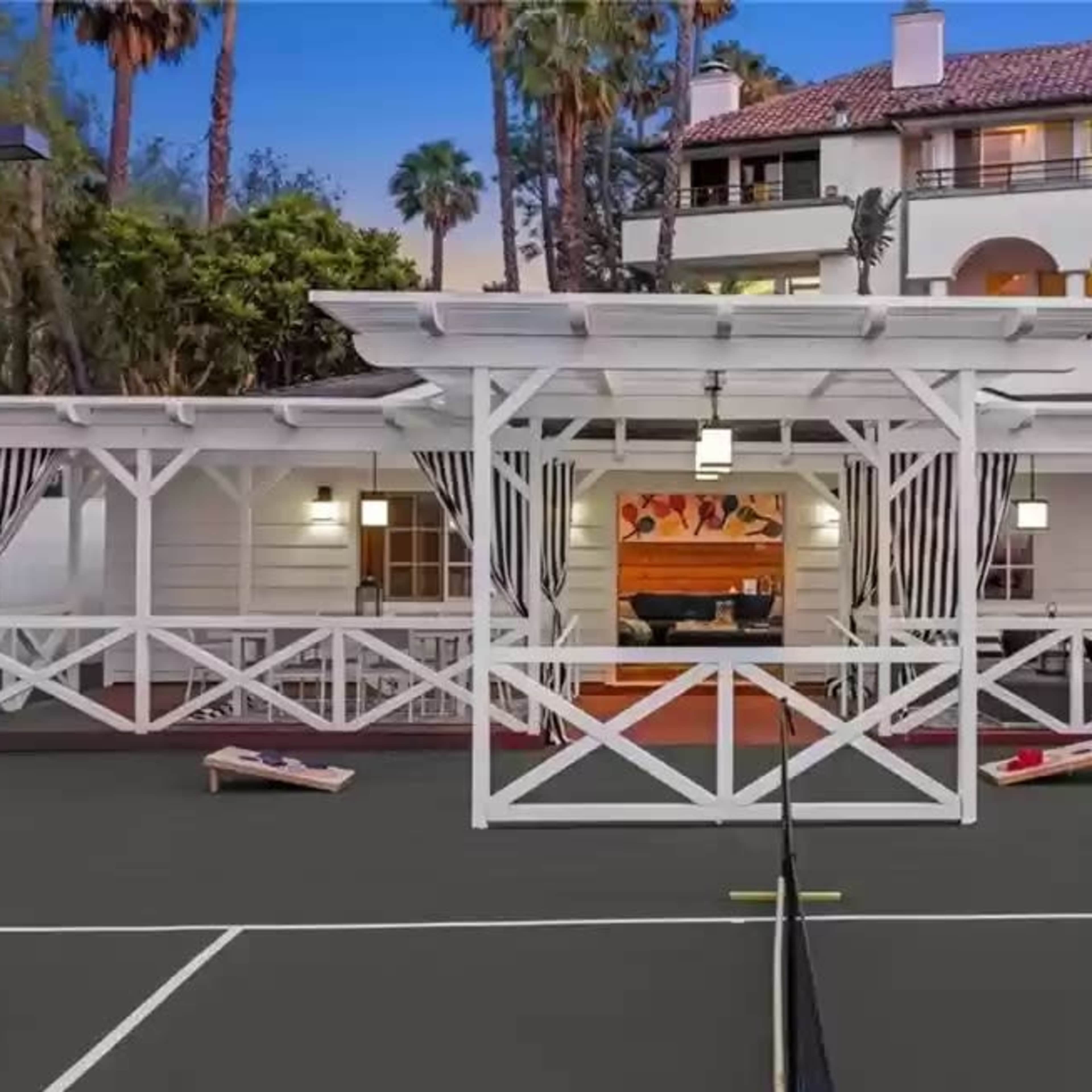 A tennis court with a white gazebo and a spacious multi-level building in the background surrounded by palm trees.