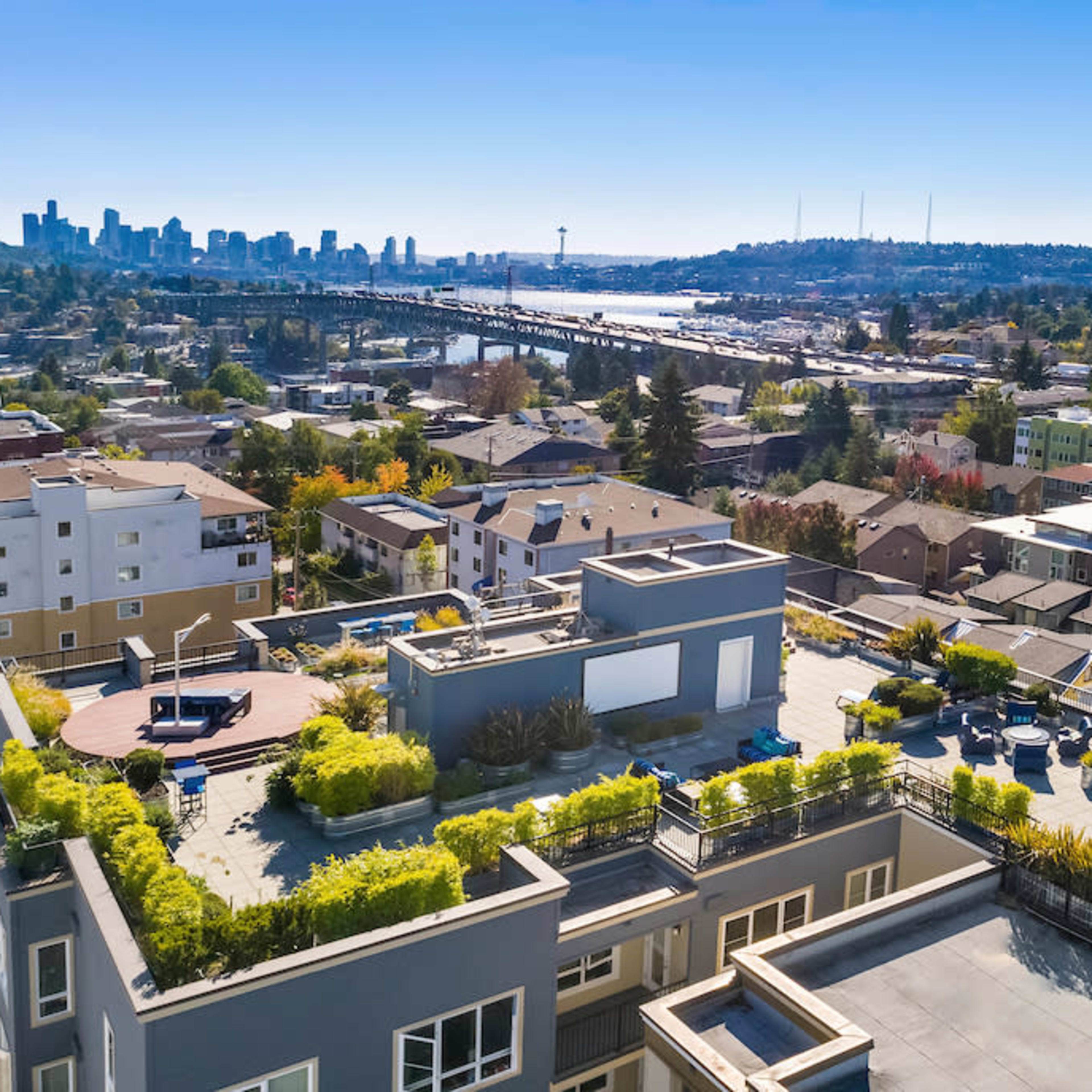 An aerial view of a residential area with modern buildings and a rooftop deck, overlooking a city skyline and a bridge across a body of water.