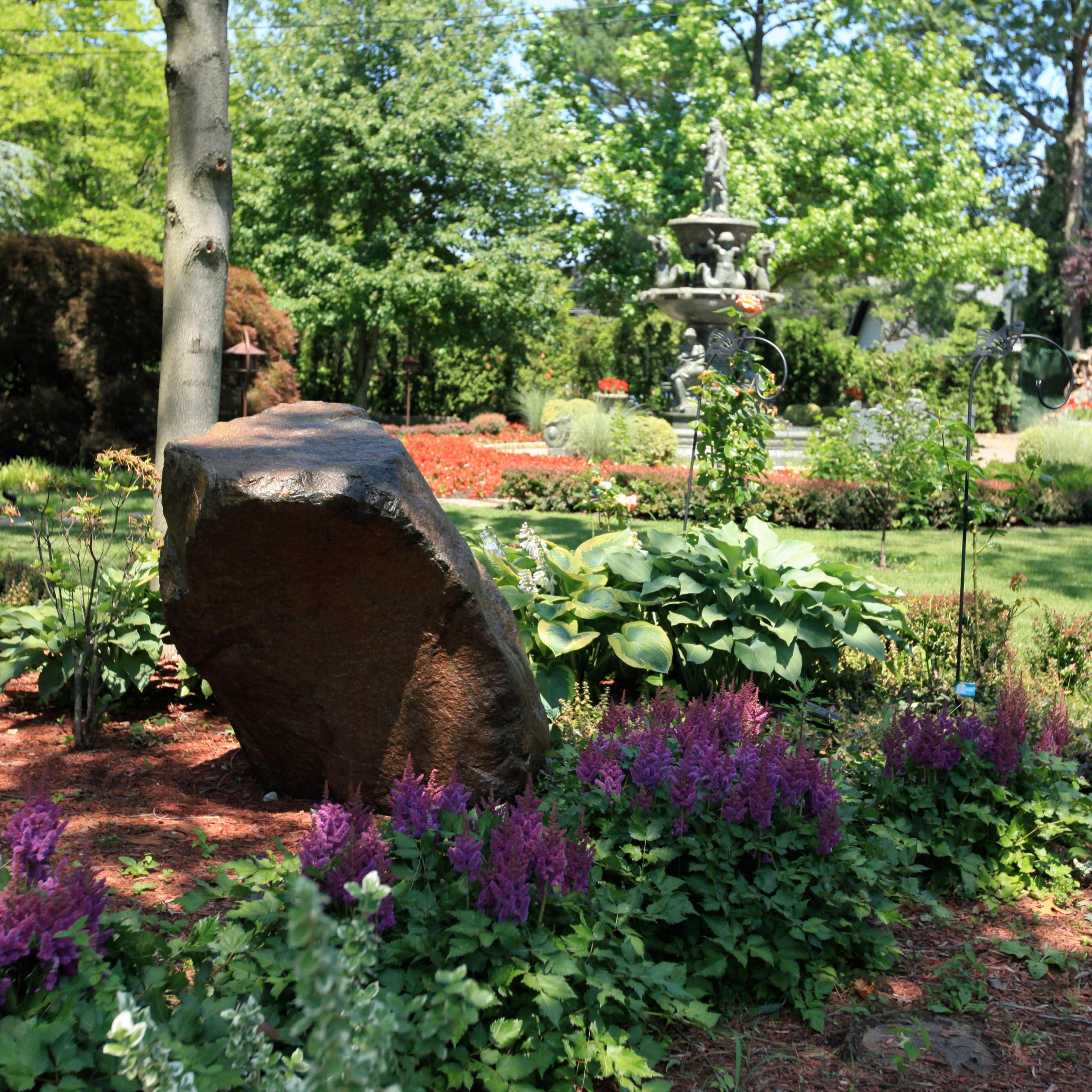 A large rock is positioned among colorful flowering plants in a well-maintained garden featuring a fountain in the background.
