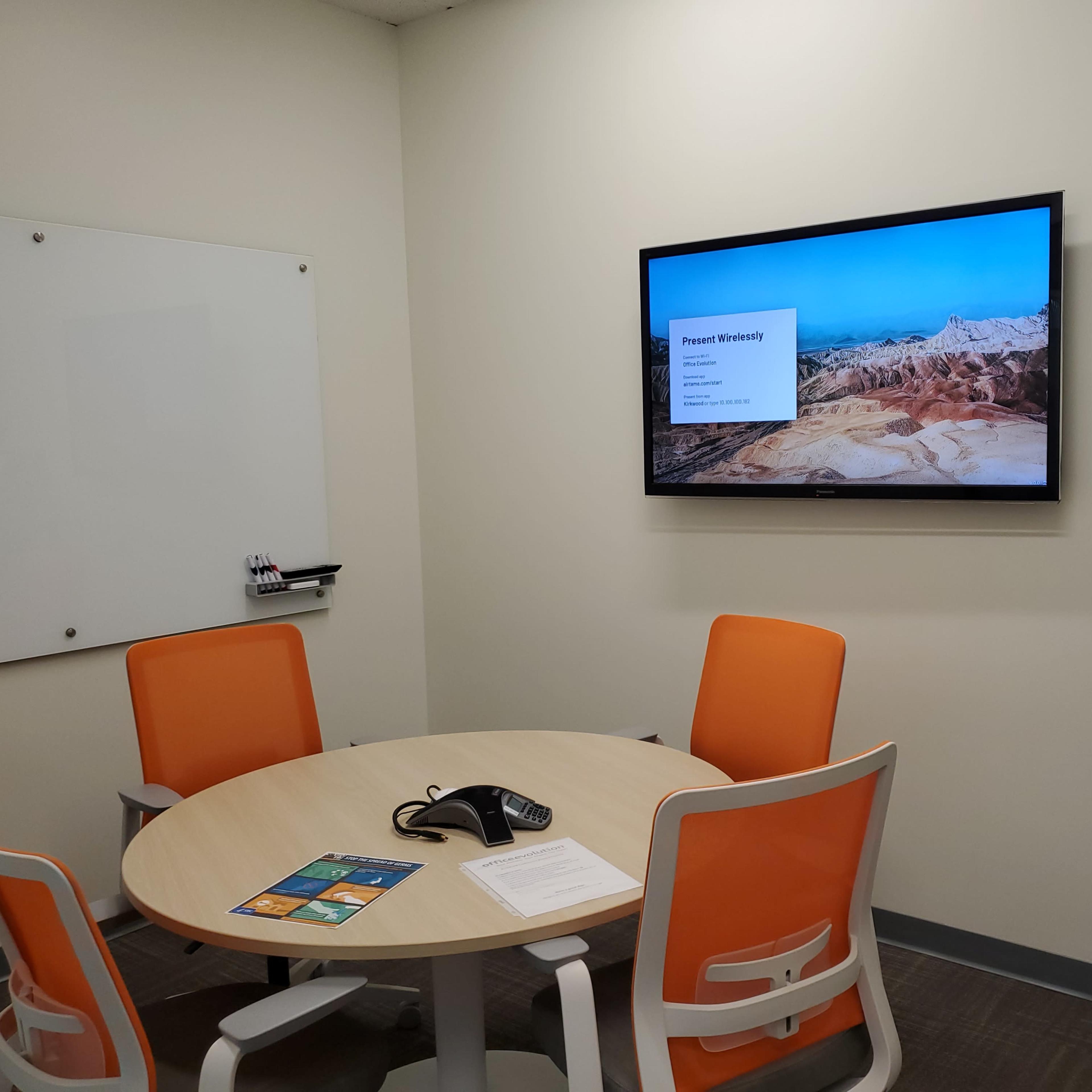 A small conference room features a round table with four orange chairs, a telephone, and a wall-mounted screen displaying a presentation.