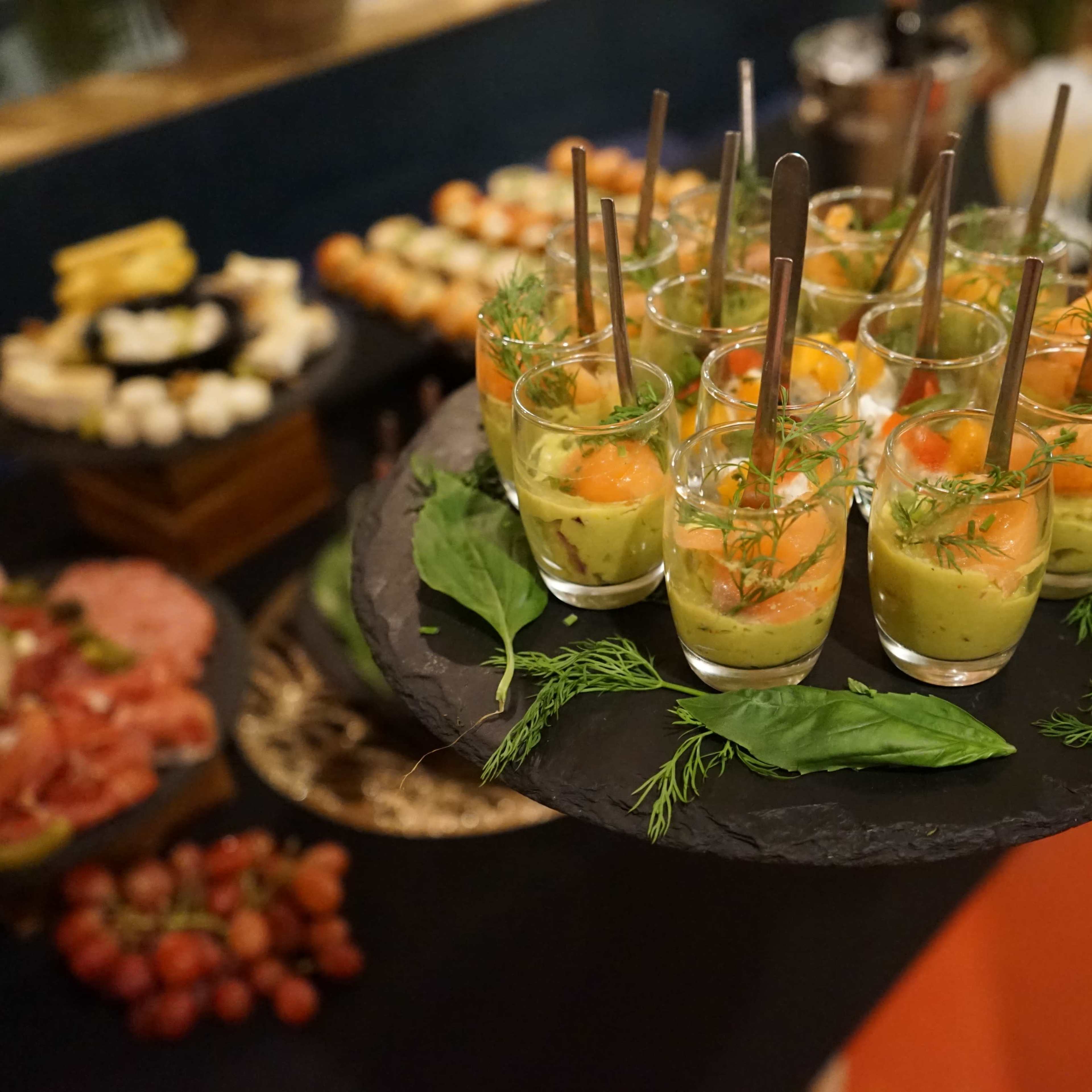 A tray holds several shot glasses filled with layered colorful appetizers, garnished with herbs and accompanied by a variety of other food items in the background.