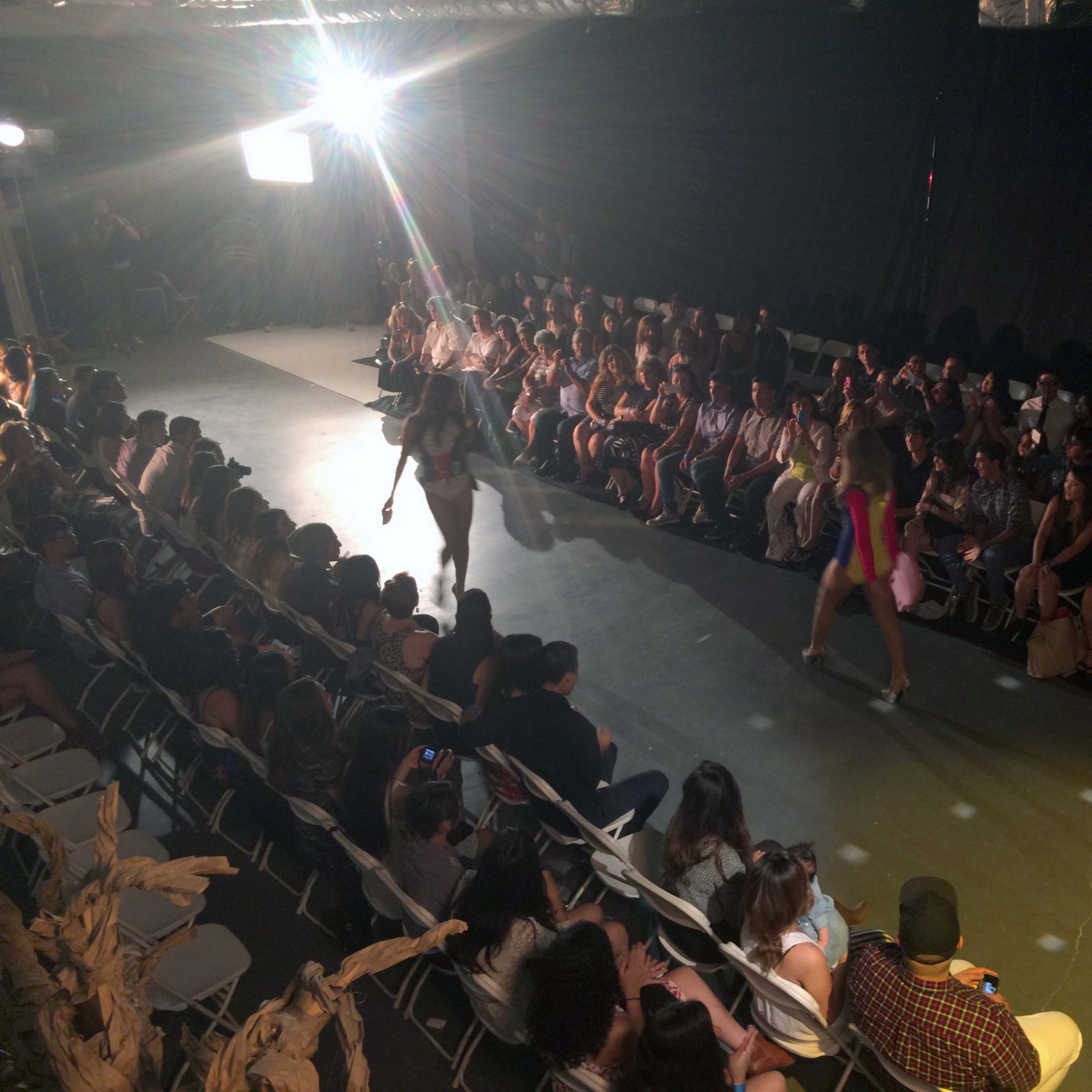 A fashion runway show is taking place with models walking down the catwalk in front of an audience seated on both sides.