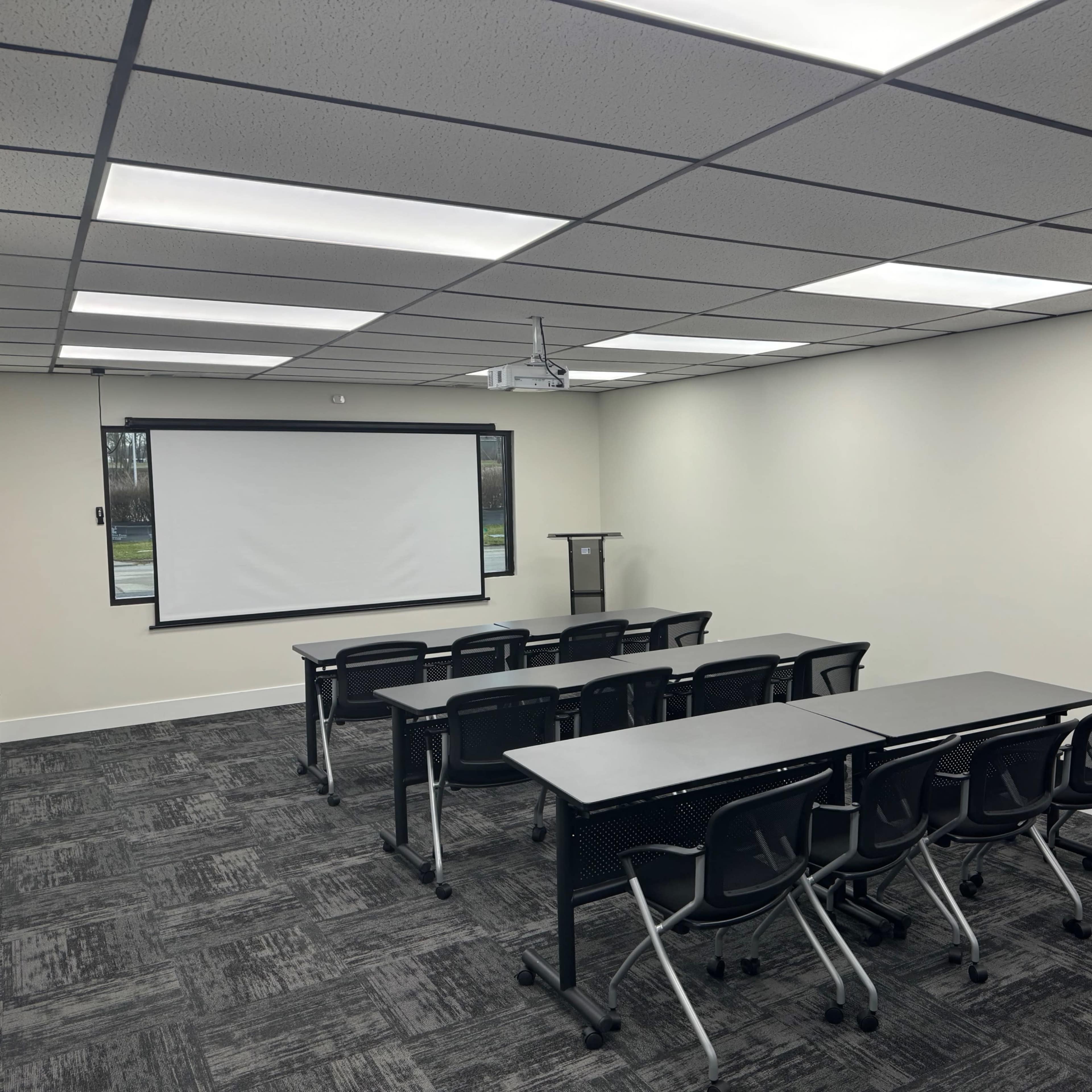 A modern, empty classroom or conference room arranged with several tables and chairs facing a projector screen at the front.