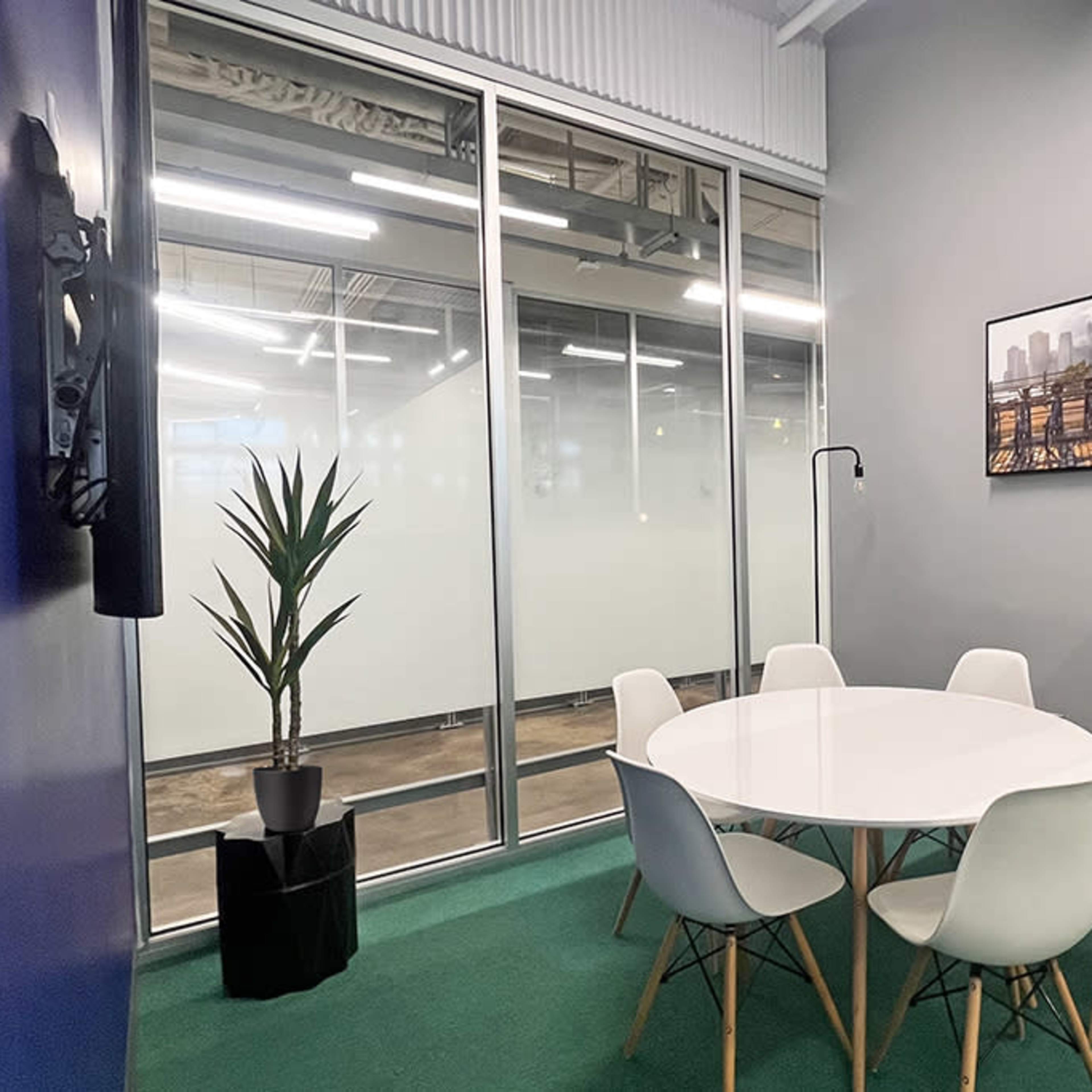 A small conference room features a round white table surrounded by six chairs, a potted plant, and a large wall-mounted television.