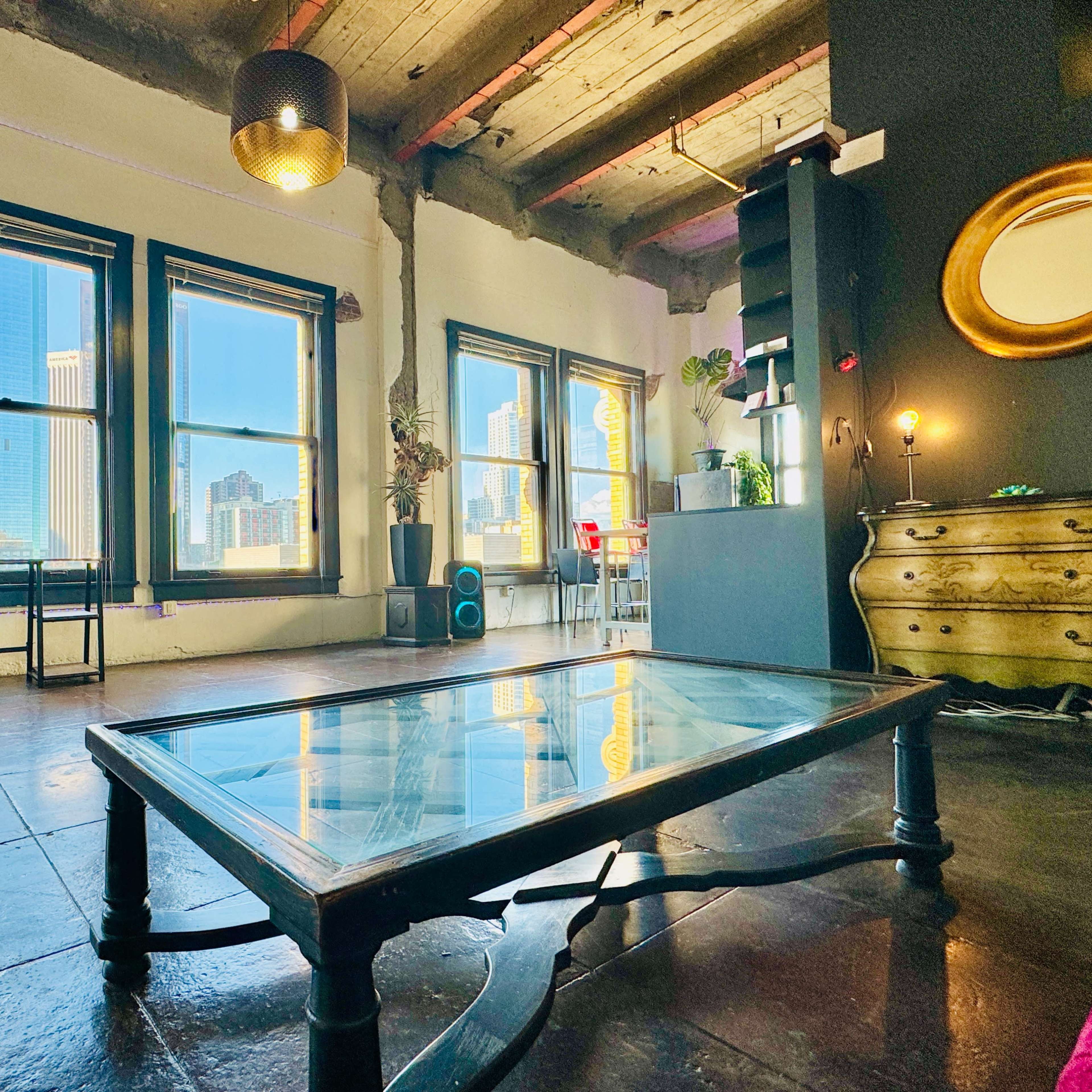 A modern, sunlit room with large windows, a glass coffee table, and various plants arranged around the space.