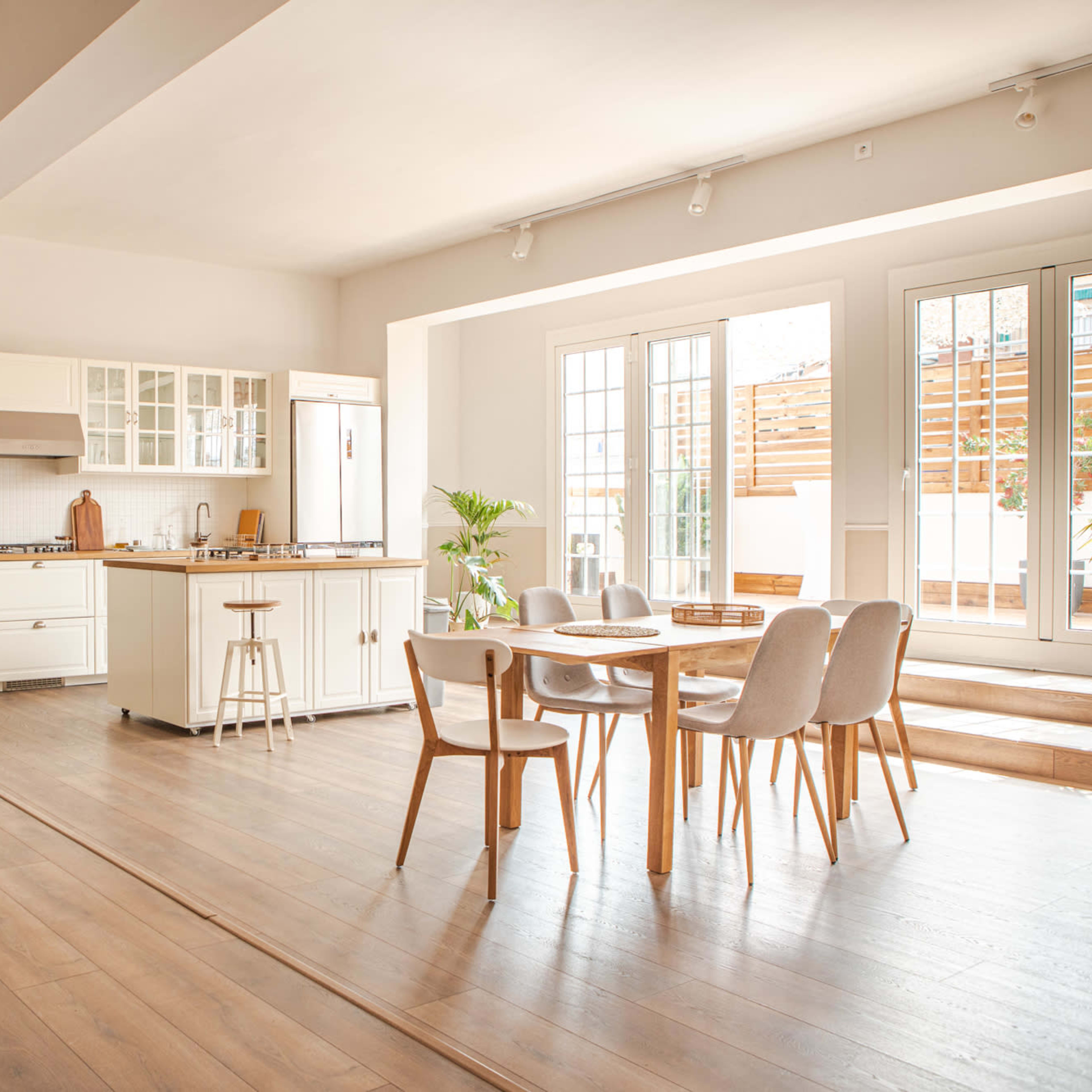 A bright kitchen features white cabinetry, a central island with stools, and a dining table surrounded by chairs, with large windows allowing natural light to illuminate the space.
