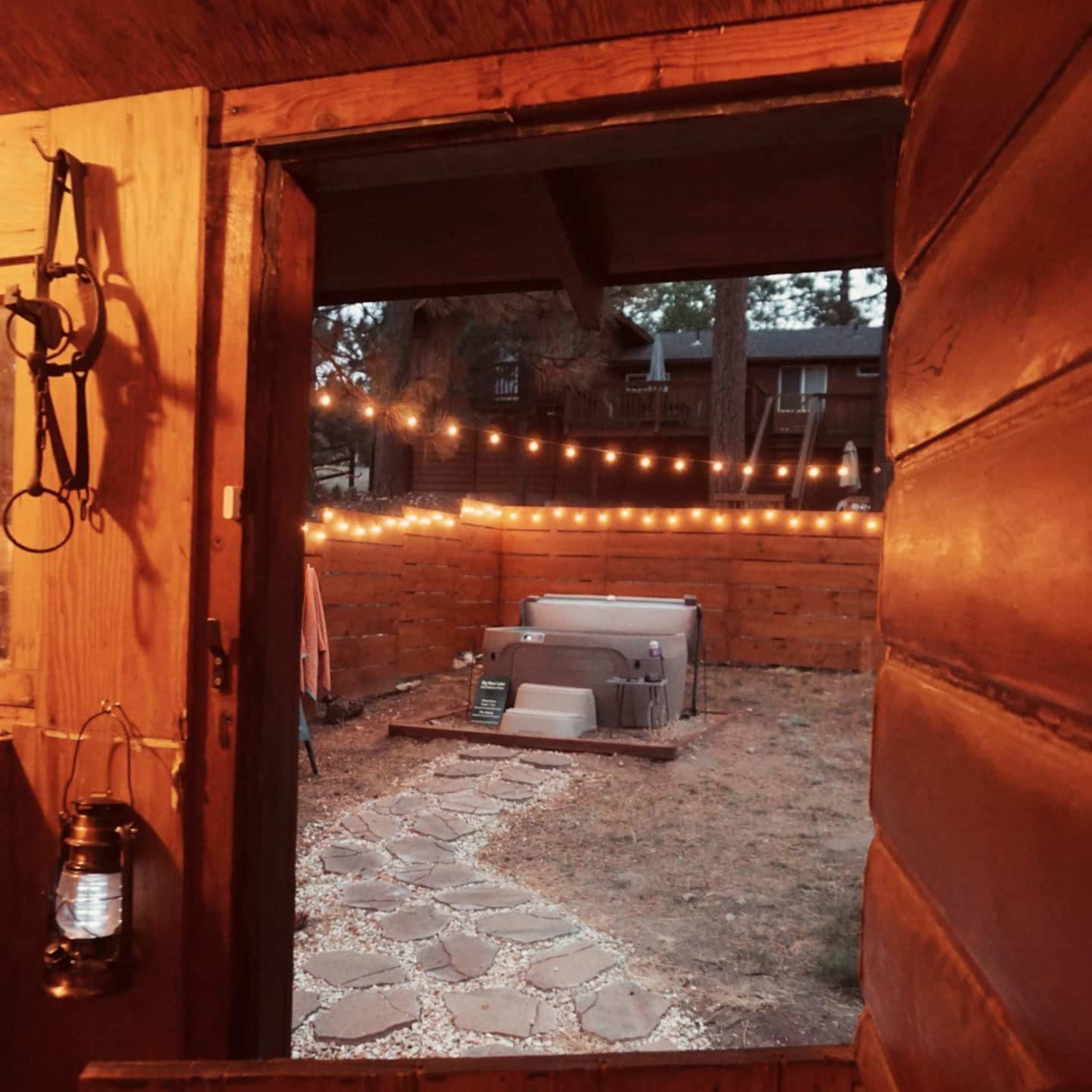 An open wooden door leading to a backyard with string lights above a hot tub surrounded by a stone path.