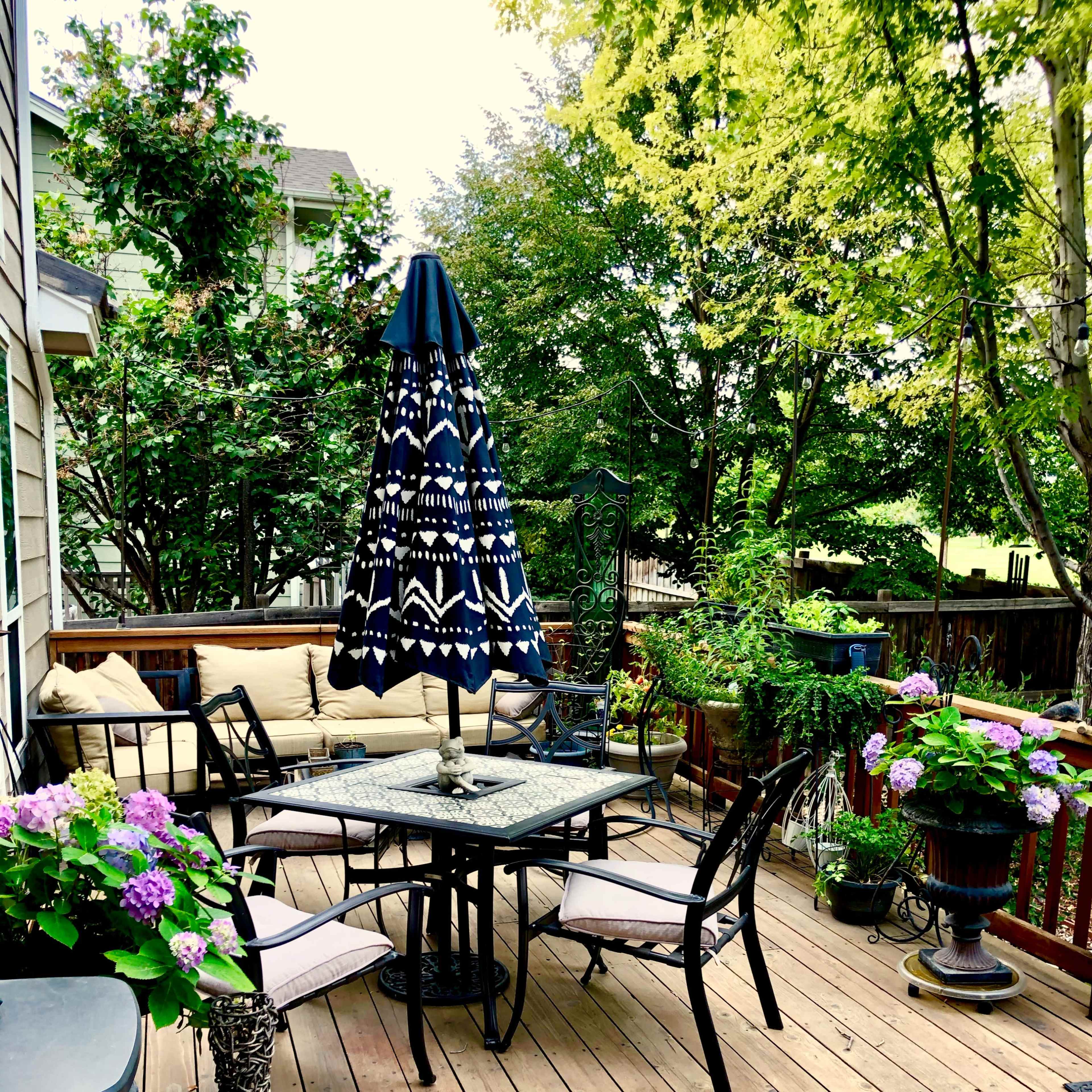 A patio setting features a round table with four chairs, an umbrella, and various potted plants in a wooden deck area.