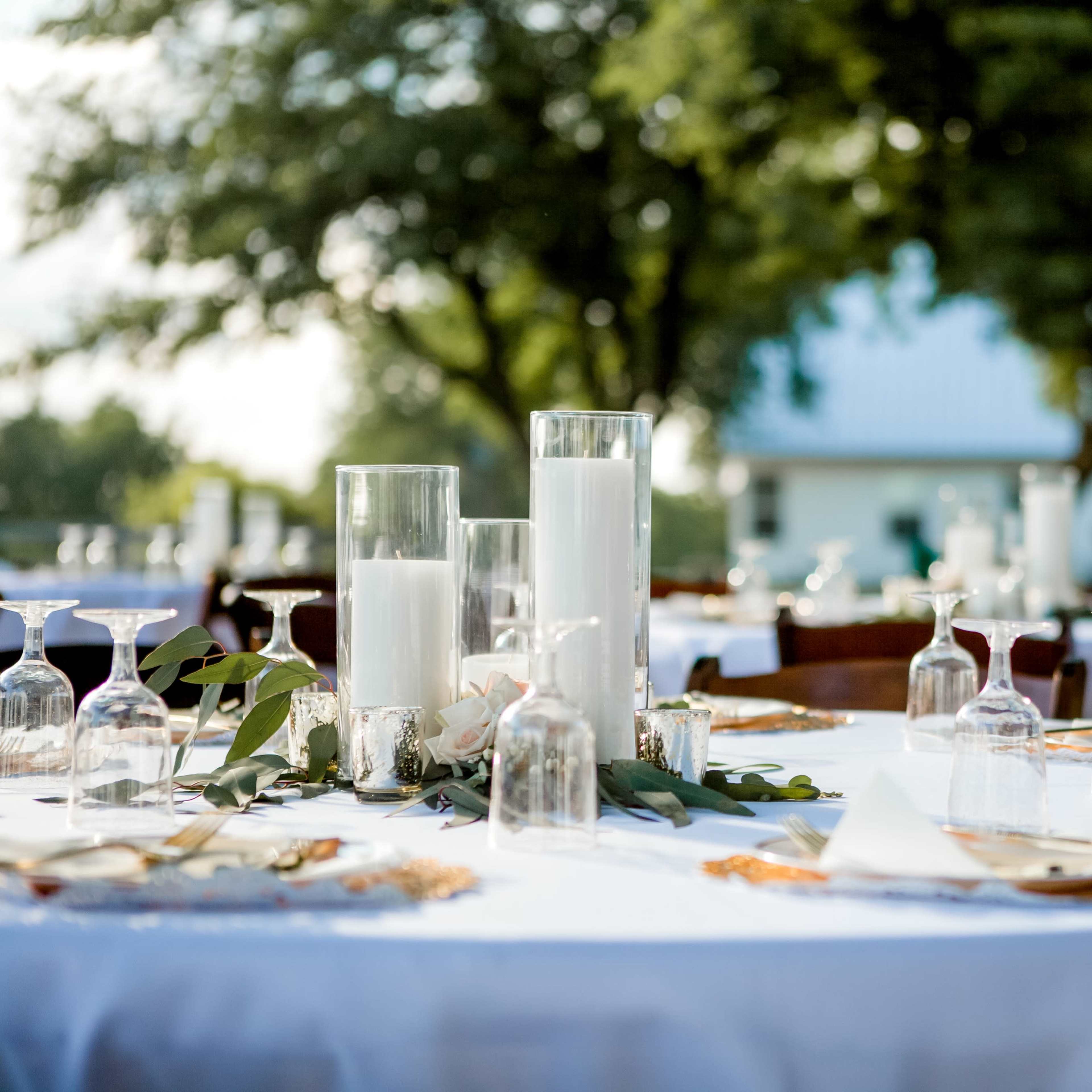 A well-set outdoor dining table features tall candles, decorative glassware, and elegant place settings surrounded by greenery.