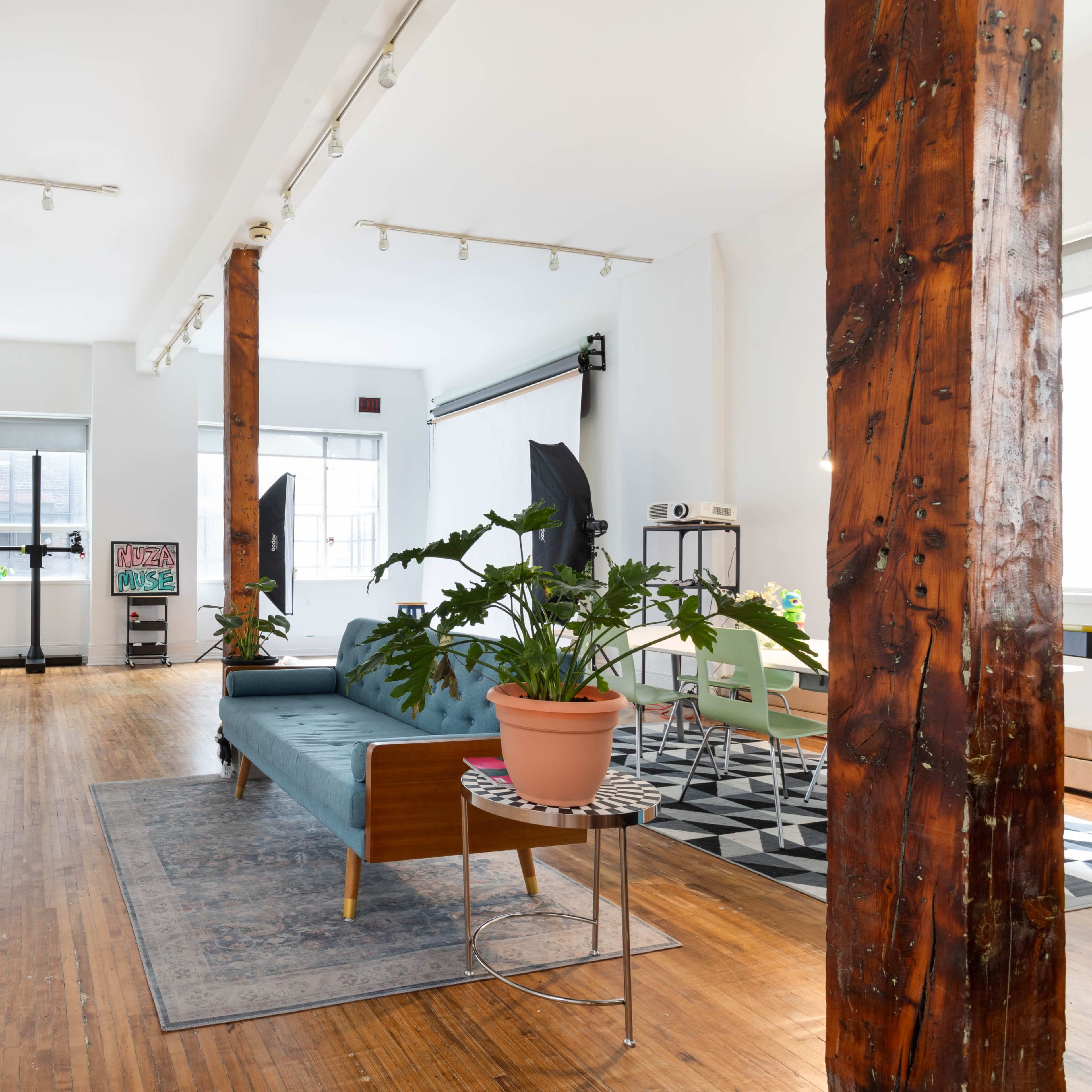 A spacious, well-lit room features wooden pillars, a blue sofa, a potted plant on a small table, and various furniture arrangements.