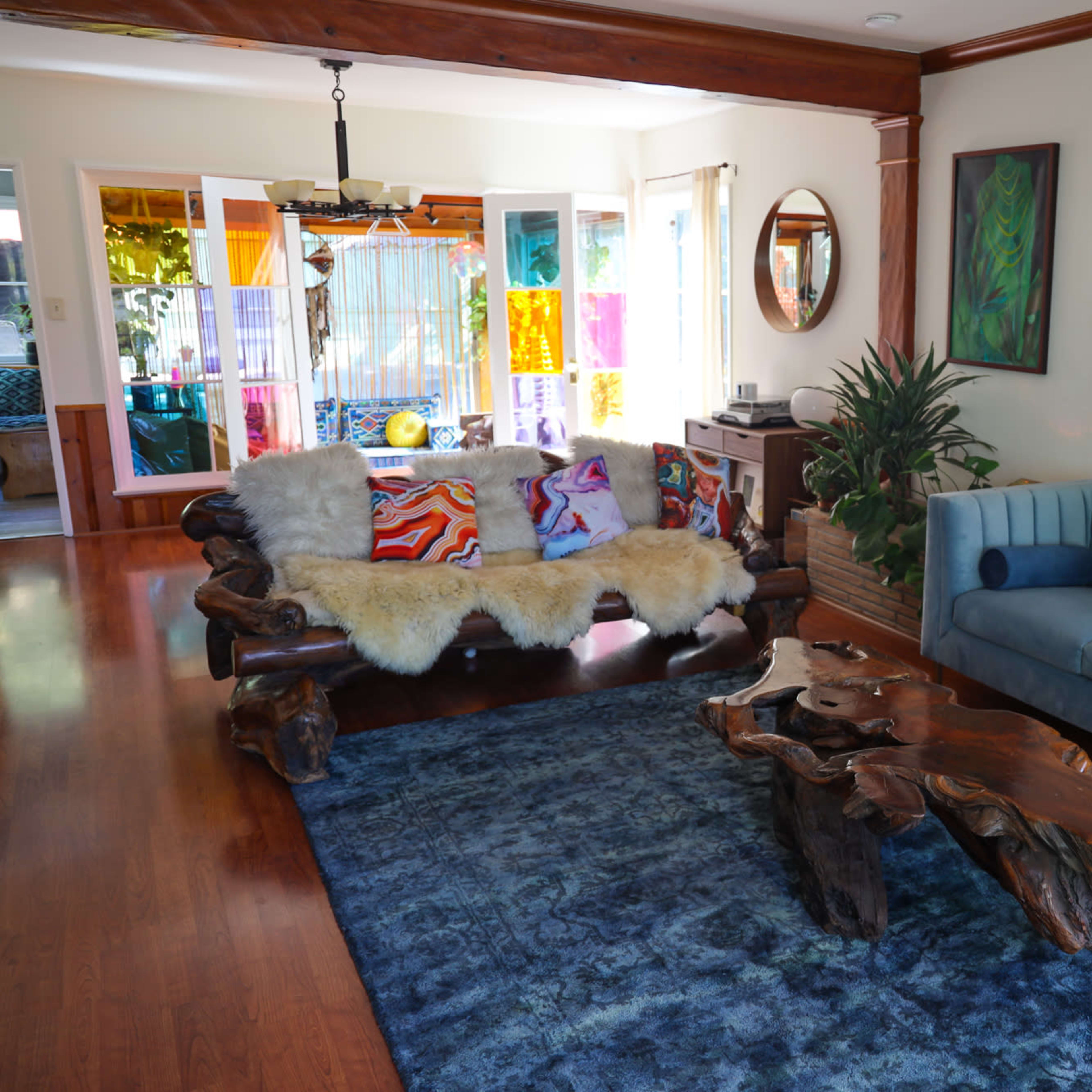 The living room features a wooden coffee table, a blue couch, a colorful patterned sofa adorned with cushions, and a large area rug on polished wood floors.