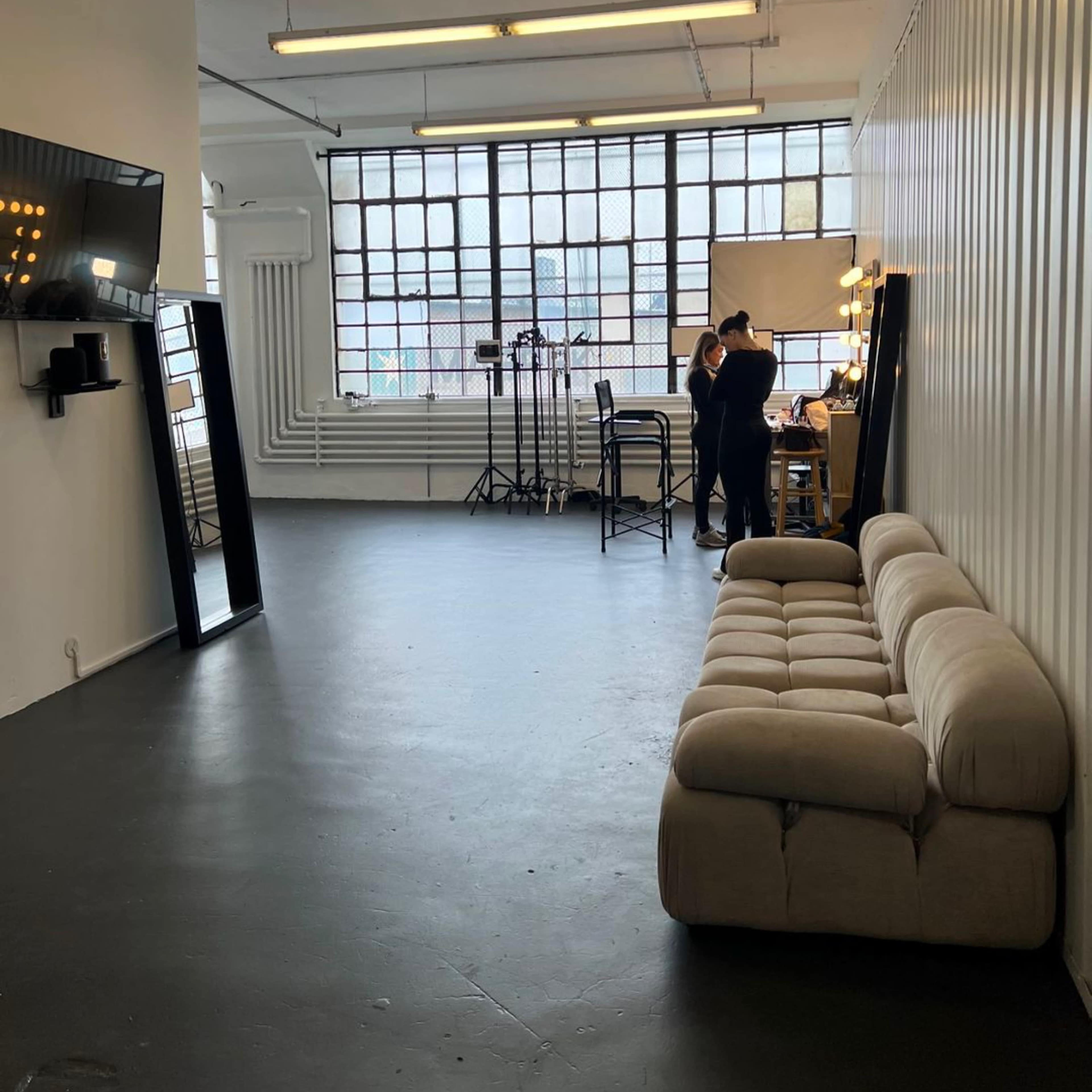 A spacious, minimalistic studio with a beige sofa, large windows, and two individuals working at a table with lighting equipment in the background.