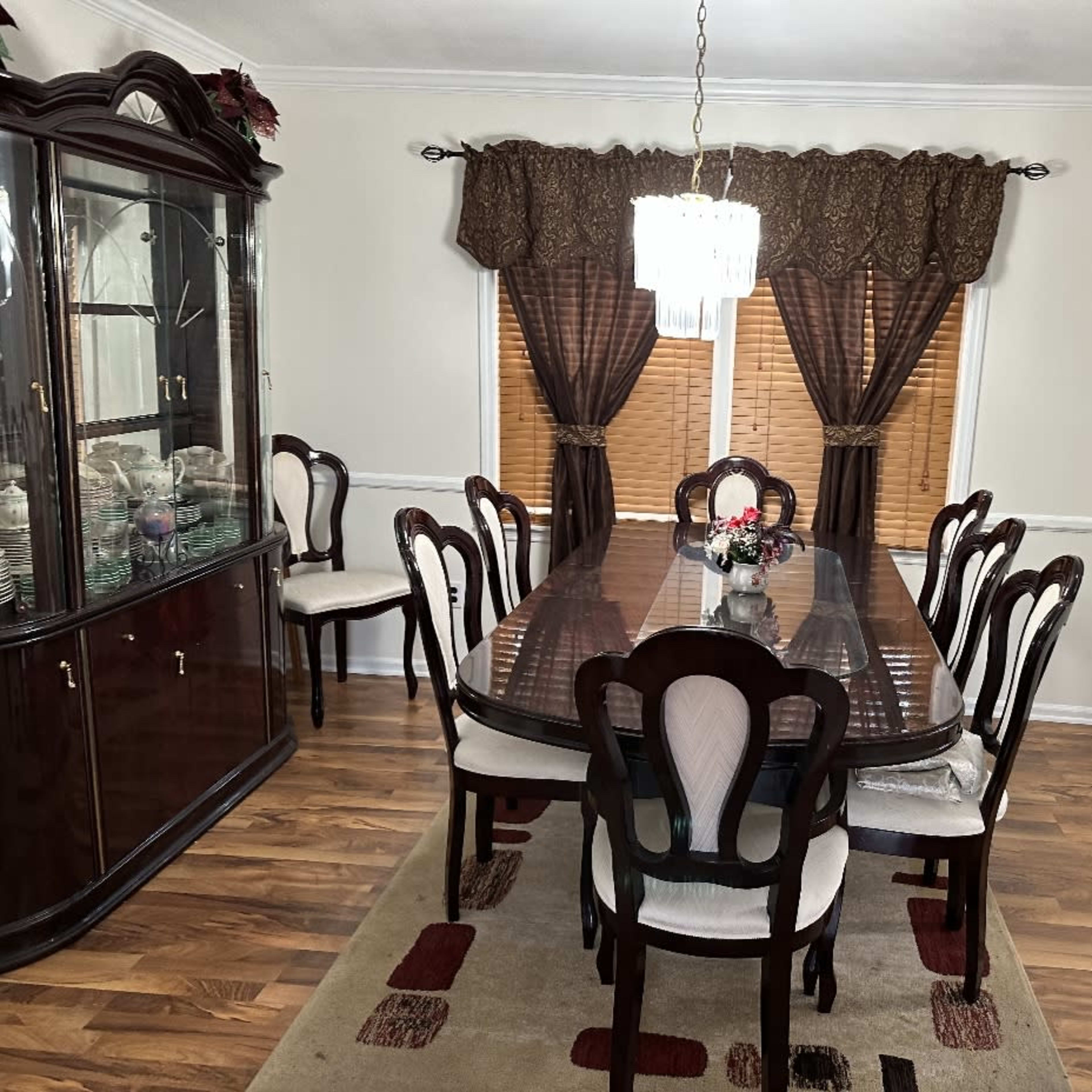 A dining room features a large table with eight chairs, a china cabinet, and windows dressed with dark curtains.
