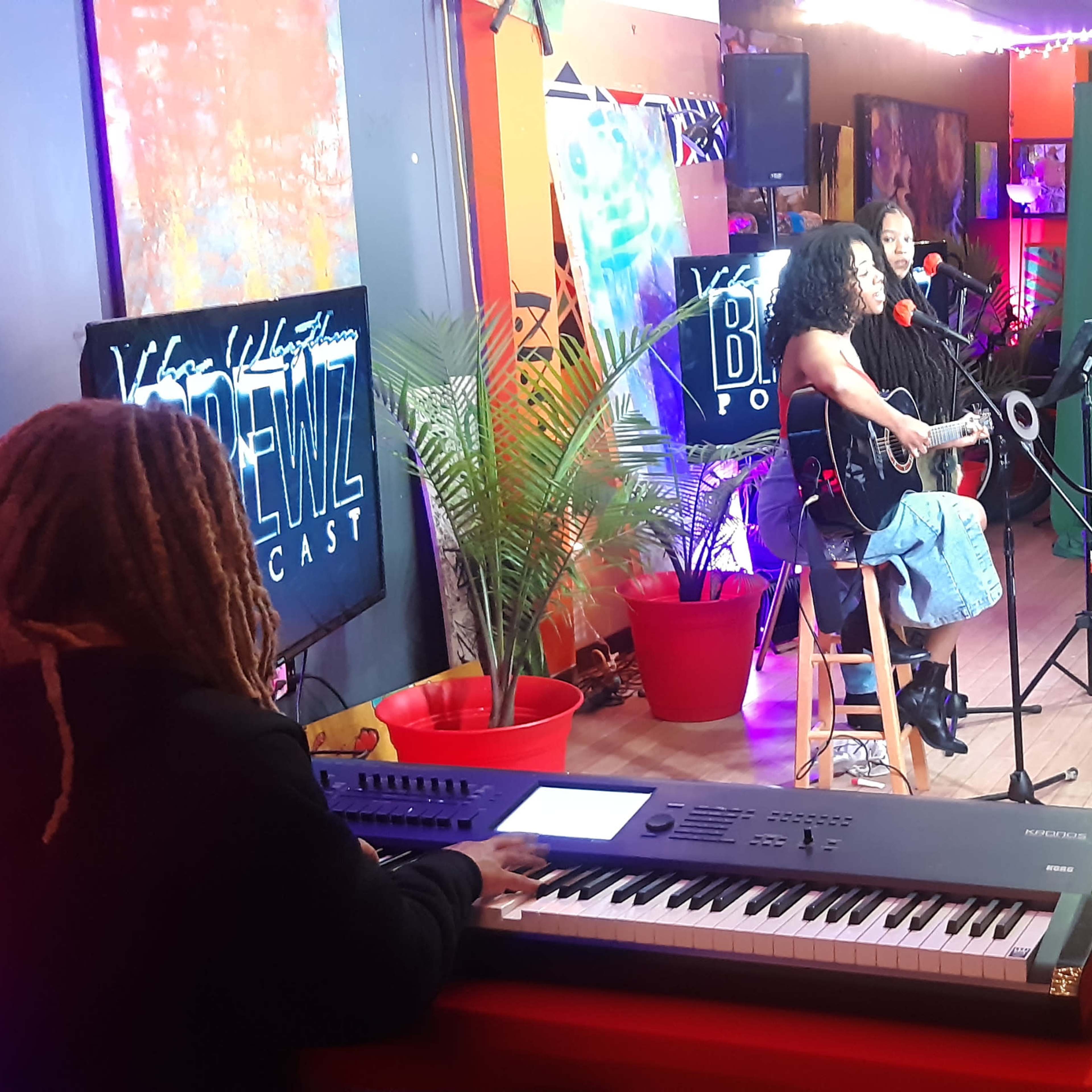 A music recording session is taking place in a brightly lit room, featuring two performers and a keyboardist, with various cameras and lighting equipment set up.