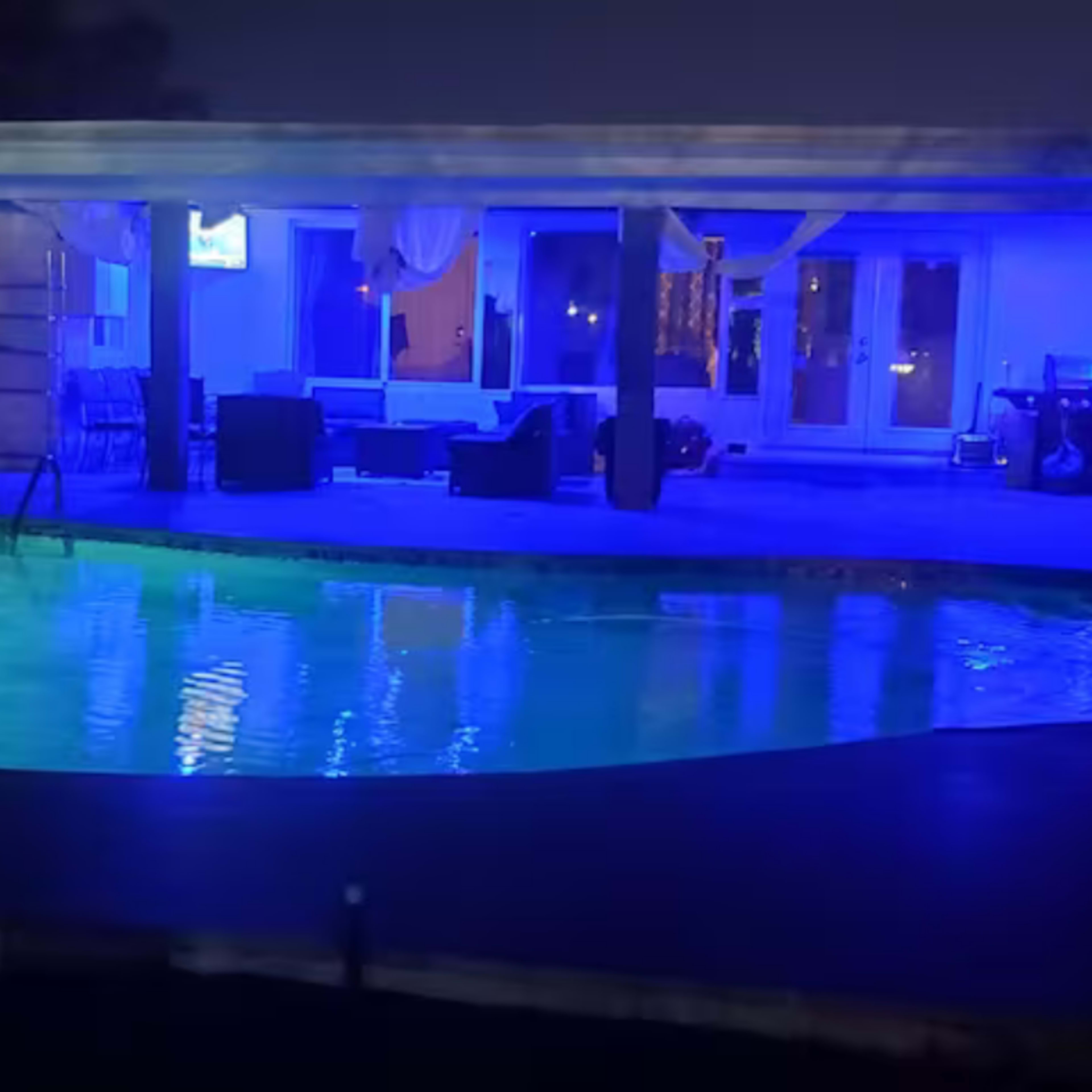 A pool with blue lighting reflects the surrounding patio area that features seating under a covered structure at night.