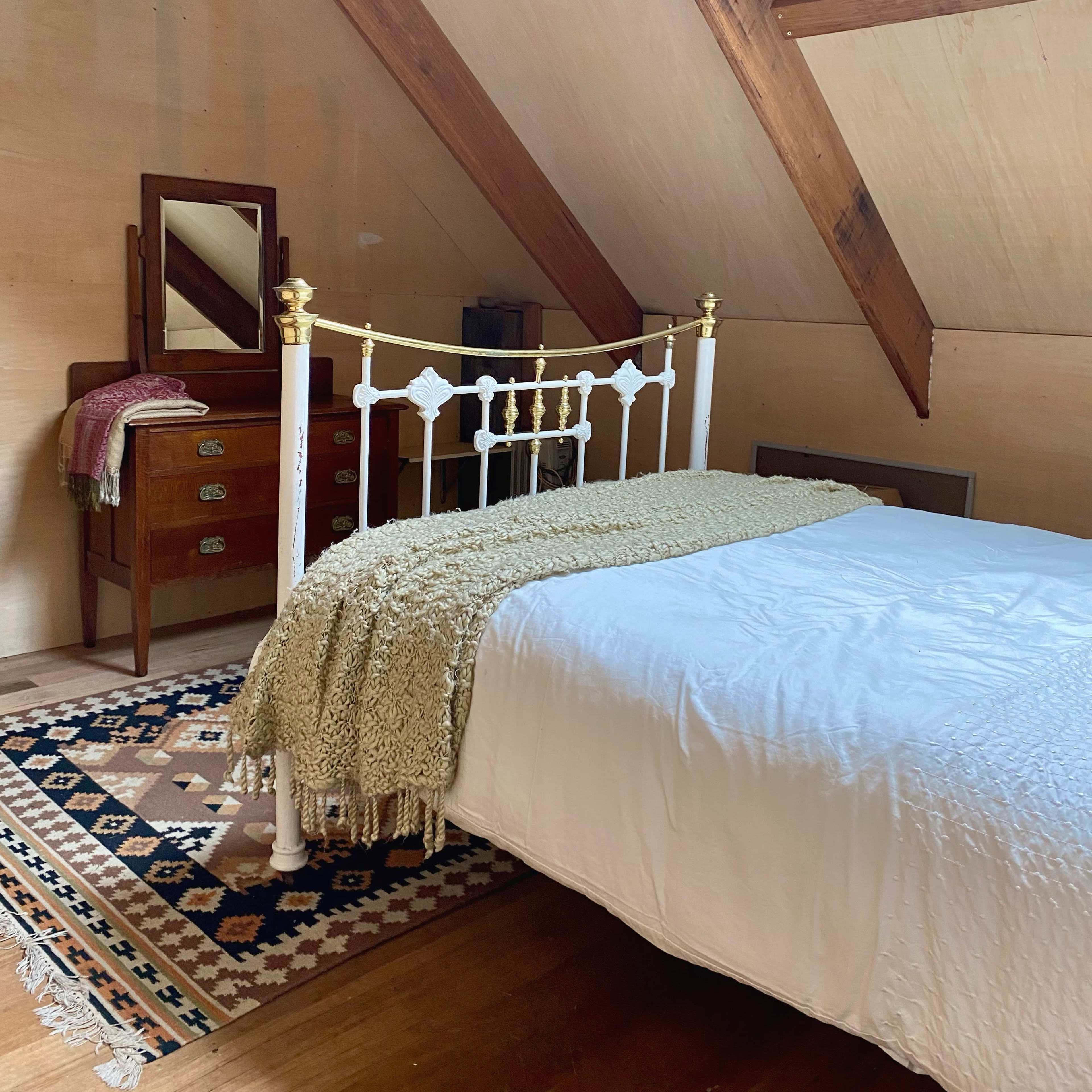 A cozy attic bedroom featuring a white metal bed with a patterned blanket, a wooden dresser, and a mirror under a slanted roof with exposed beams.