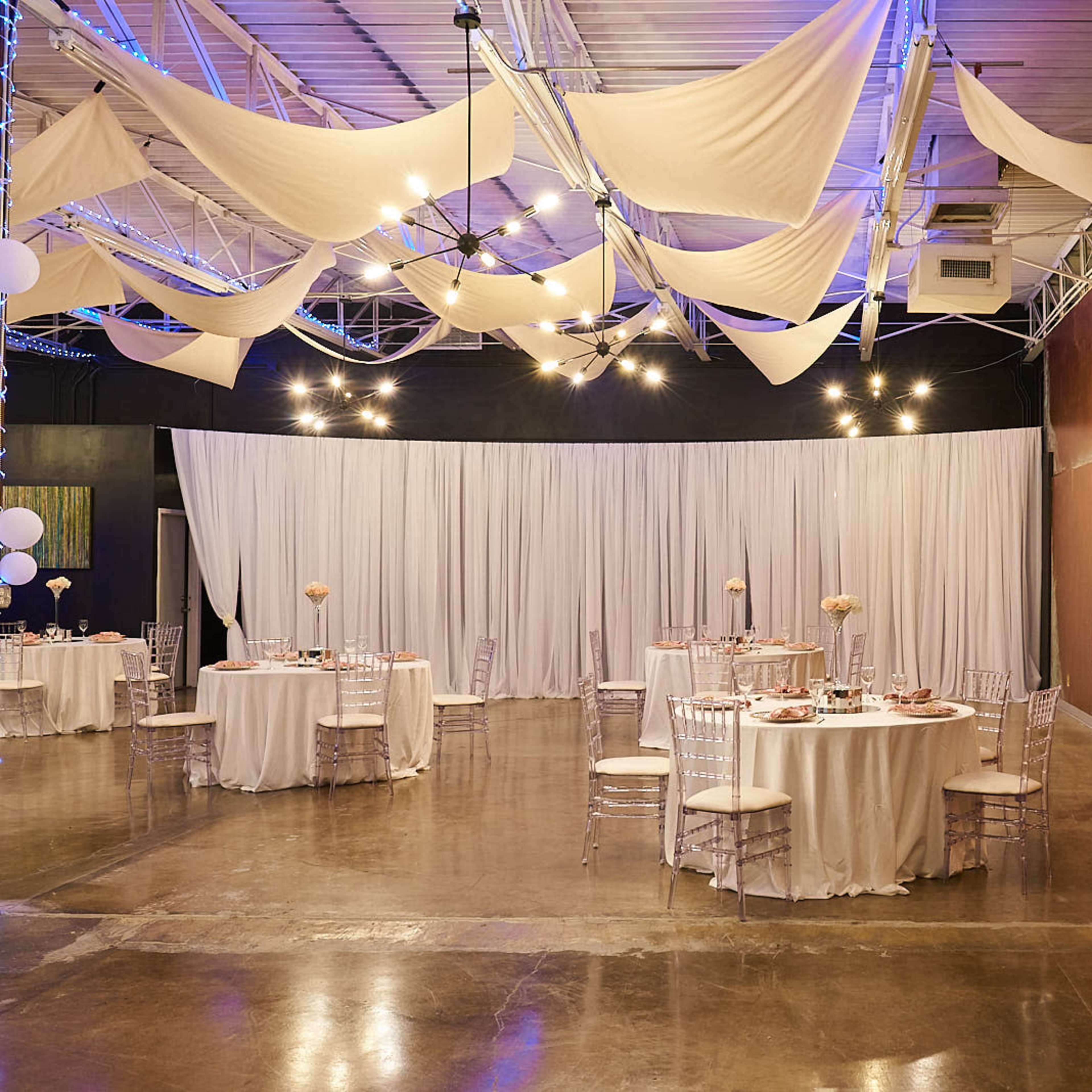 An indoor event space with round dining tables covered in white tablecloths, chair covers, and decorative centerpieces, alongside a backdrop of white drapery and modern artwork on the walls.