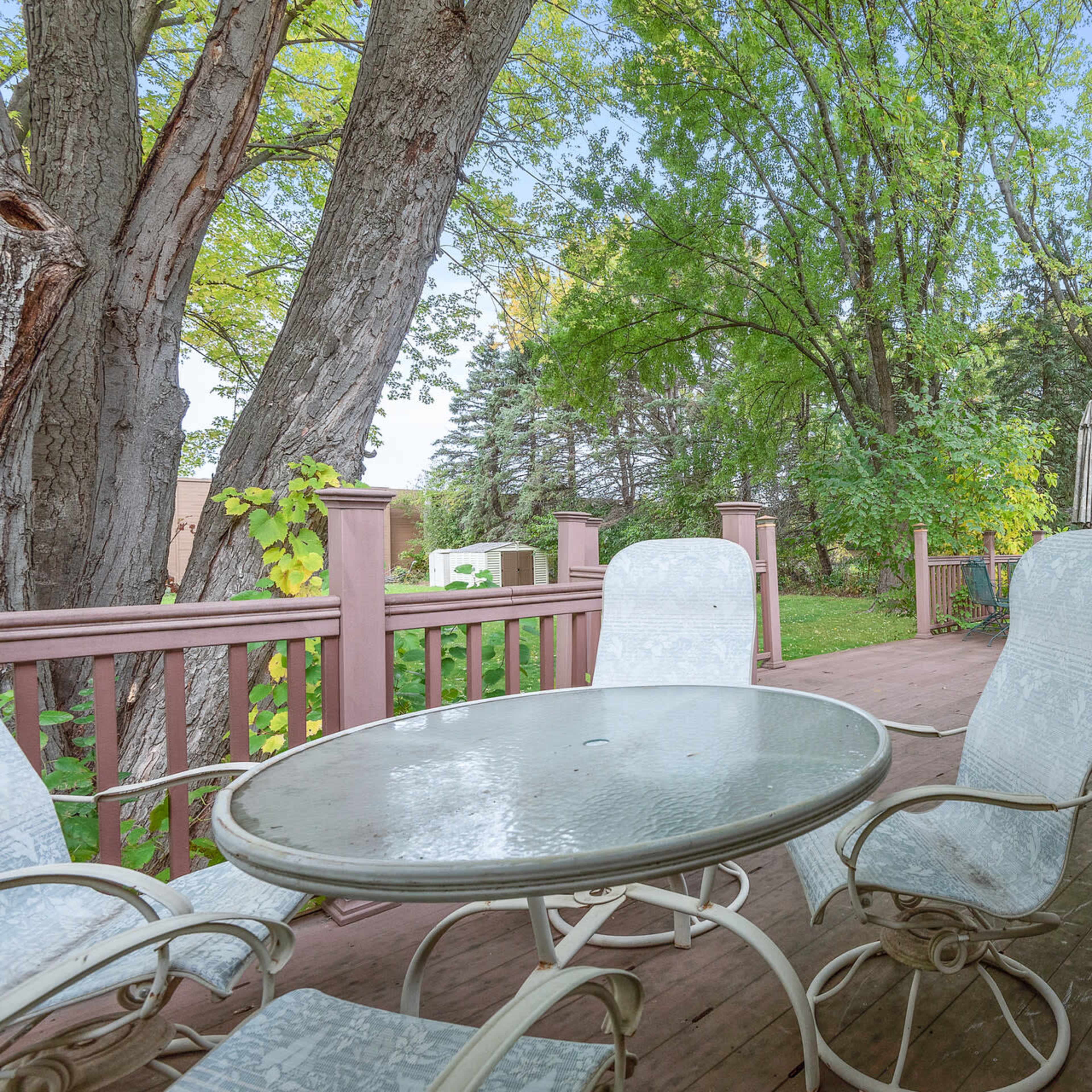 A patio area features a round table with four chairs surrounded by trees and a view of a nearby house.