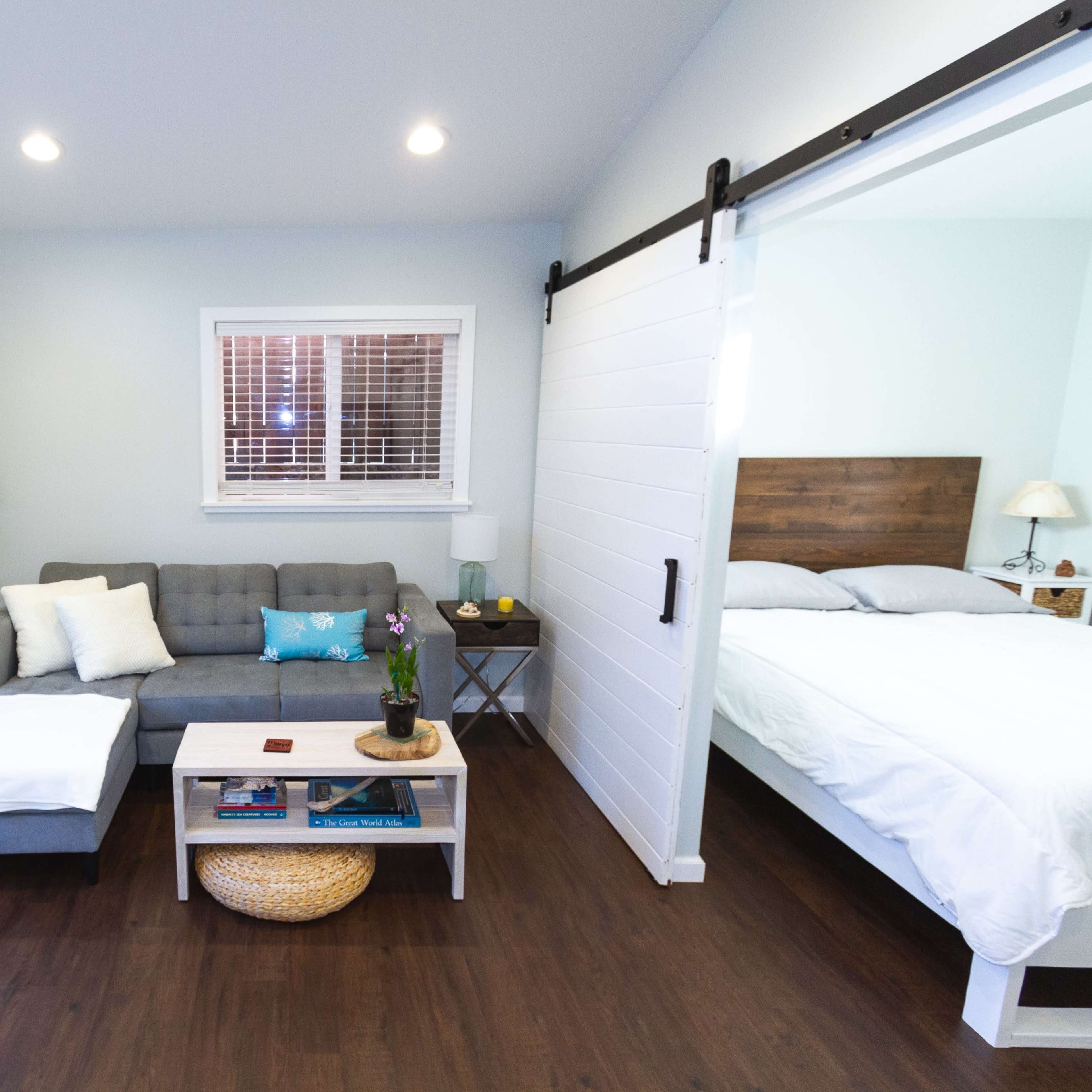 A small, well-lit living area with a gray sofa, a coffee table, and a bedroom area separated by a sliding door.