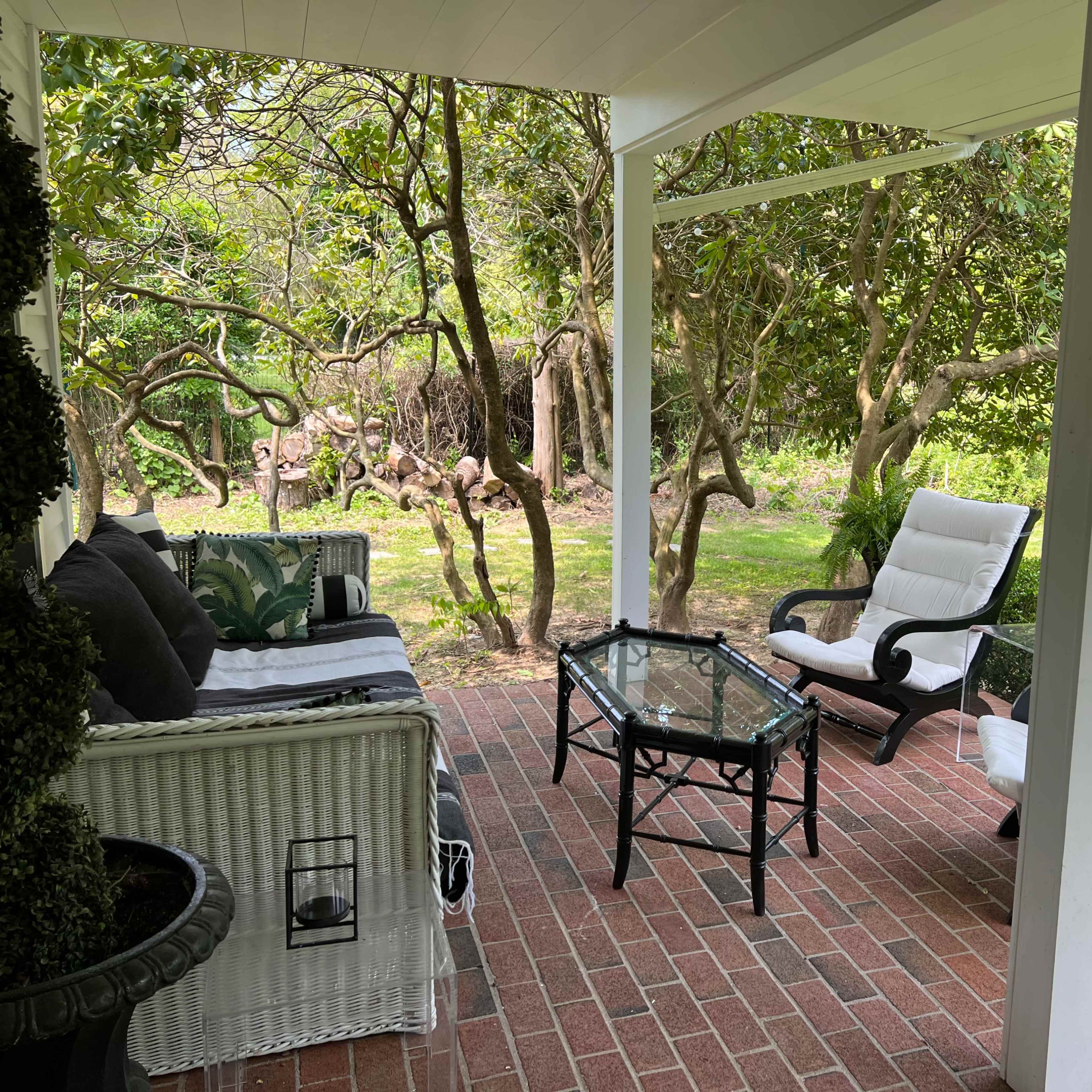 A covered patio features a glass table surrounded by two white lounge chairs and a wicker sofa, with greenery in the background.