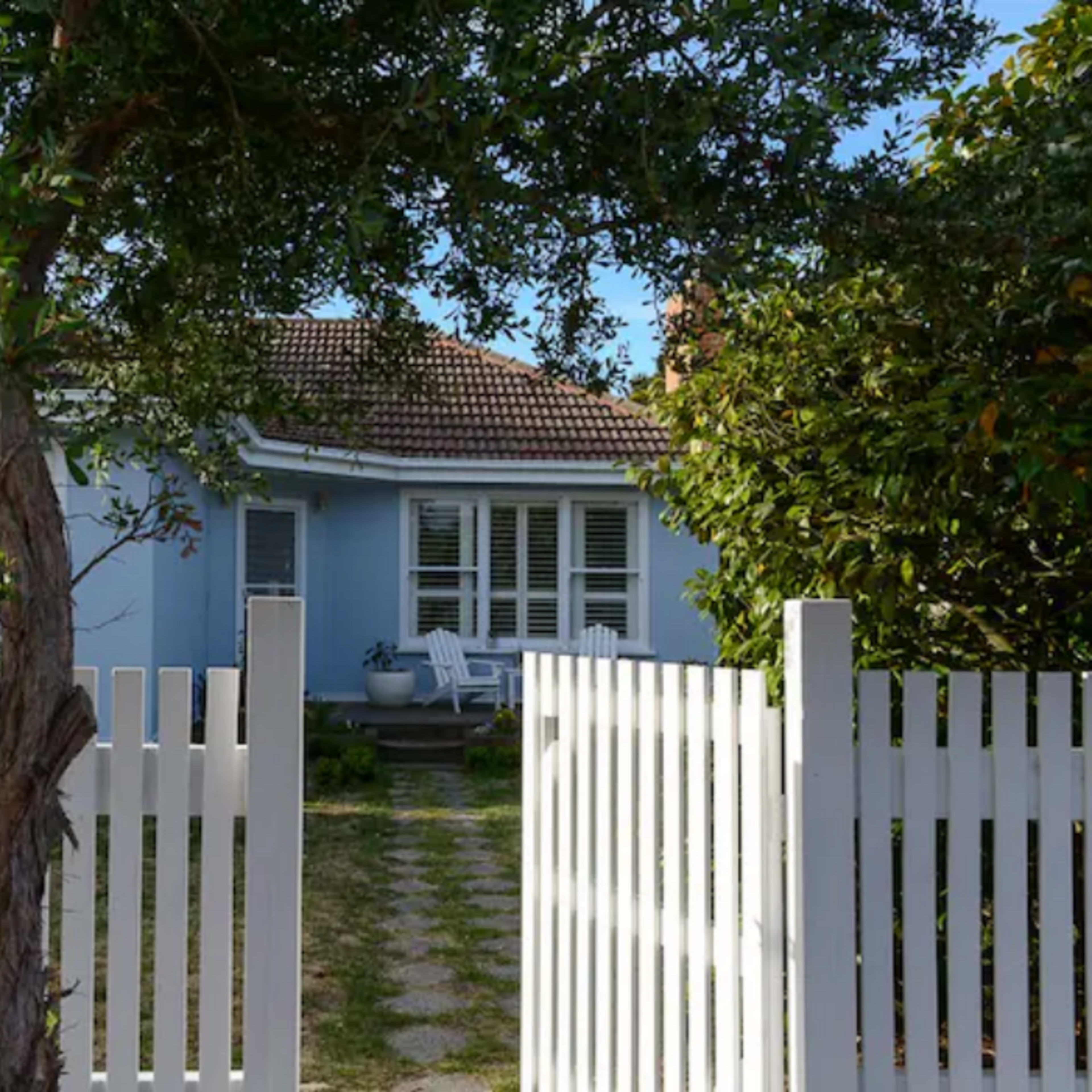 A white picket fence frames a pathway leading to a light blue house with a tiled roof and a front porch.