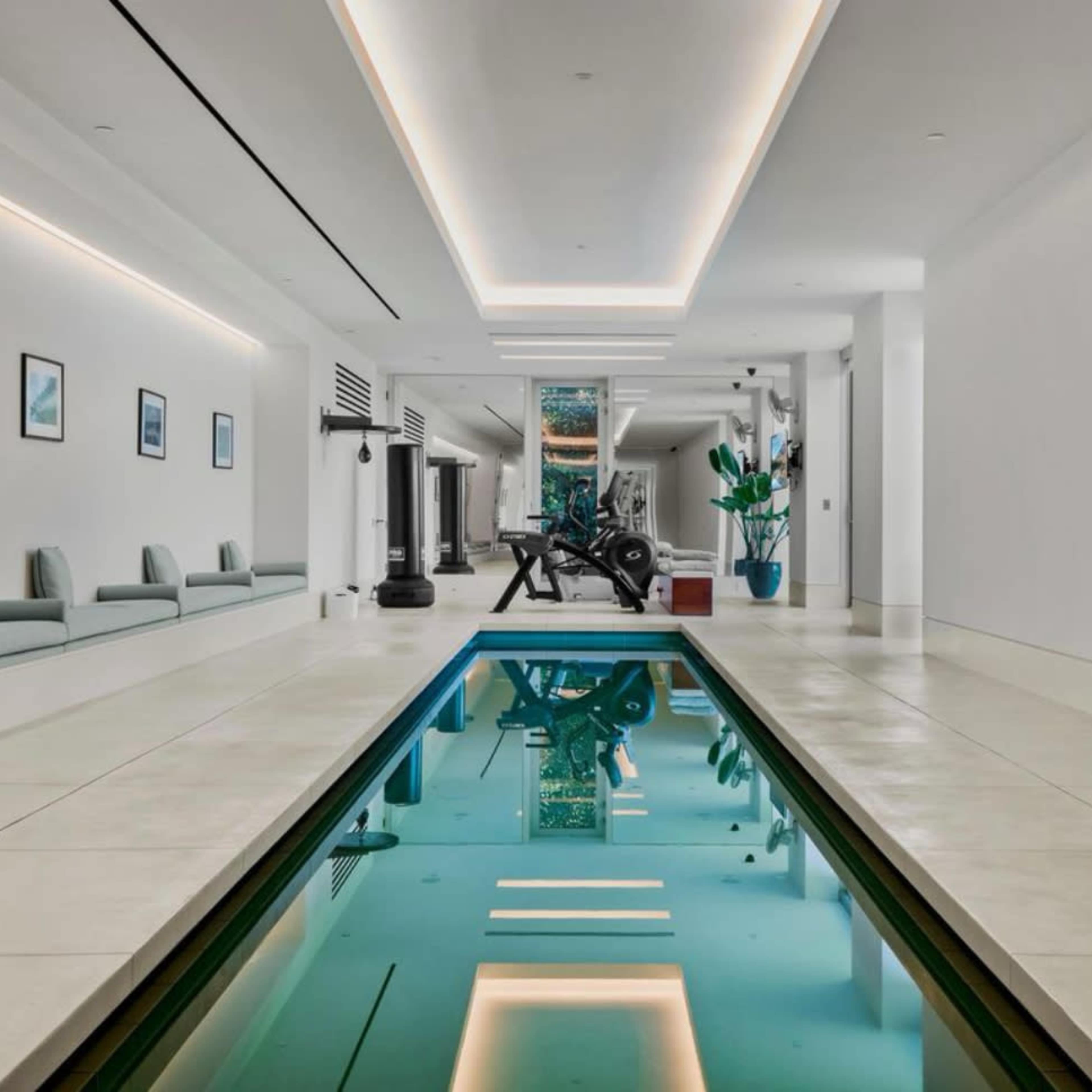 A modern indoor swimming pool surrounded by white walls, minimalist furniture, and exercise equipment in a spacious room.