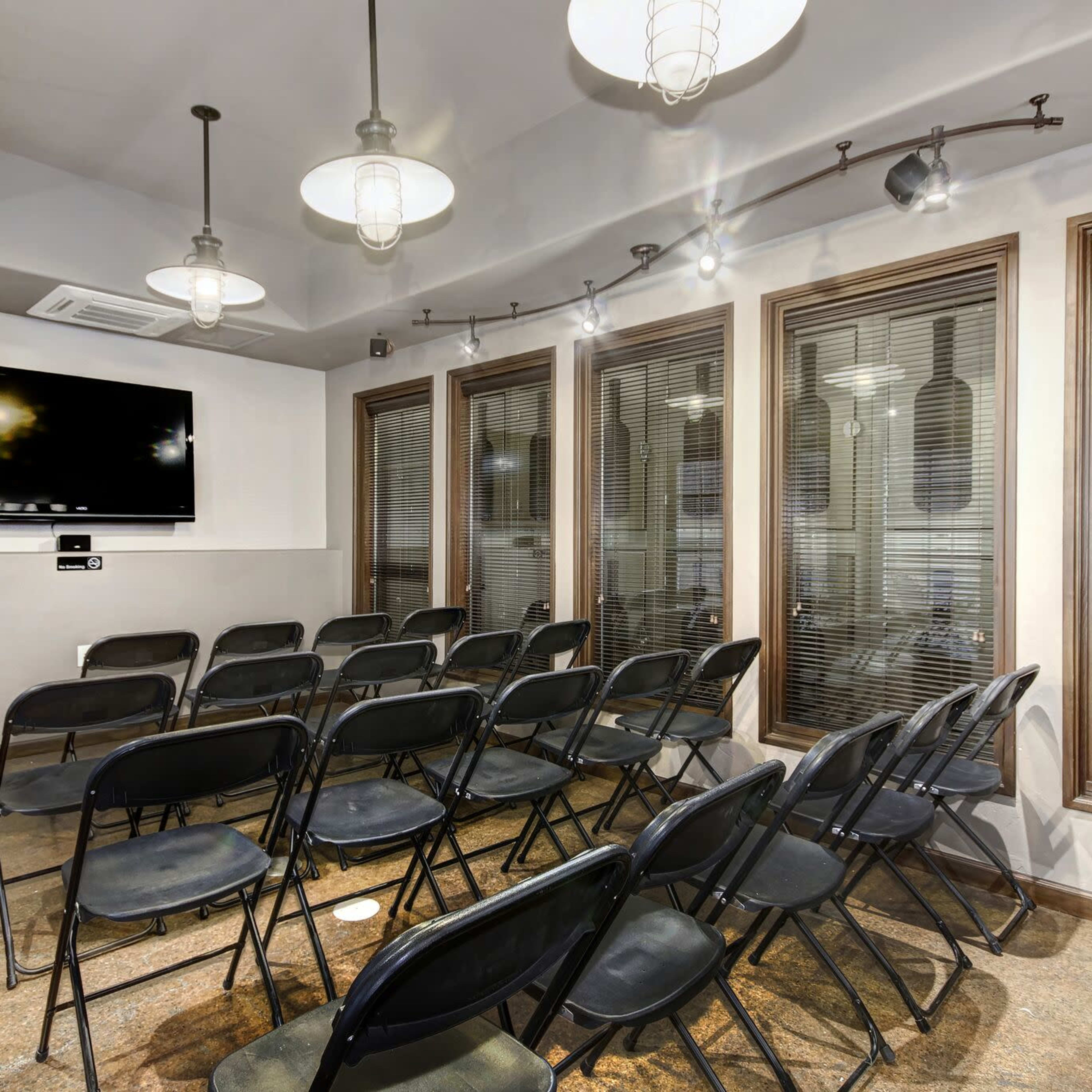 A small meeting room with several rows of black folding chairs facing a wall-mounted television and large windows with blinds.