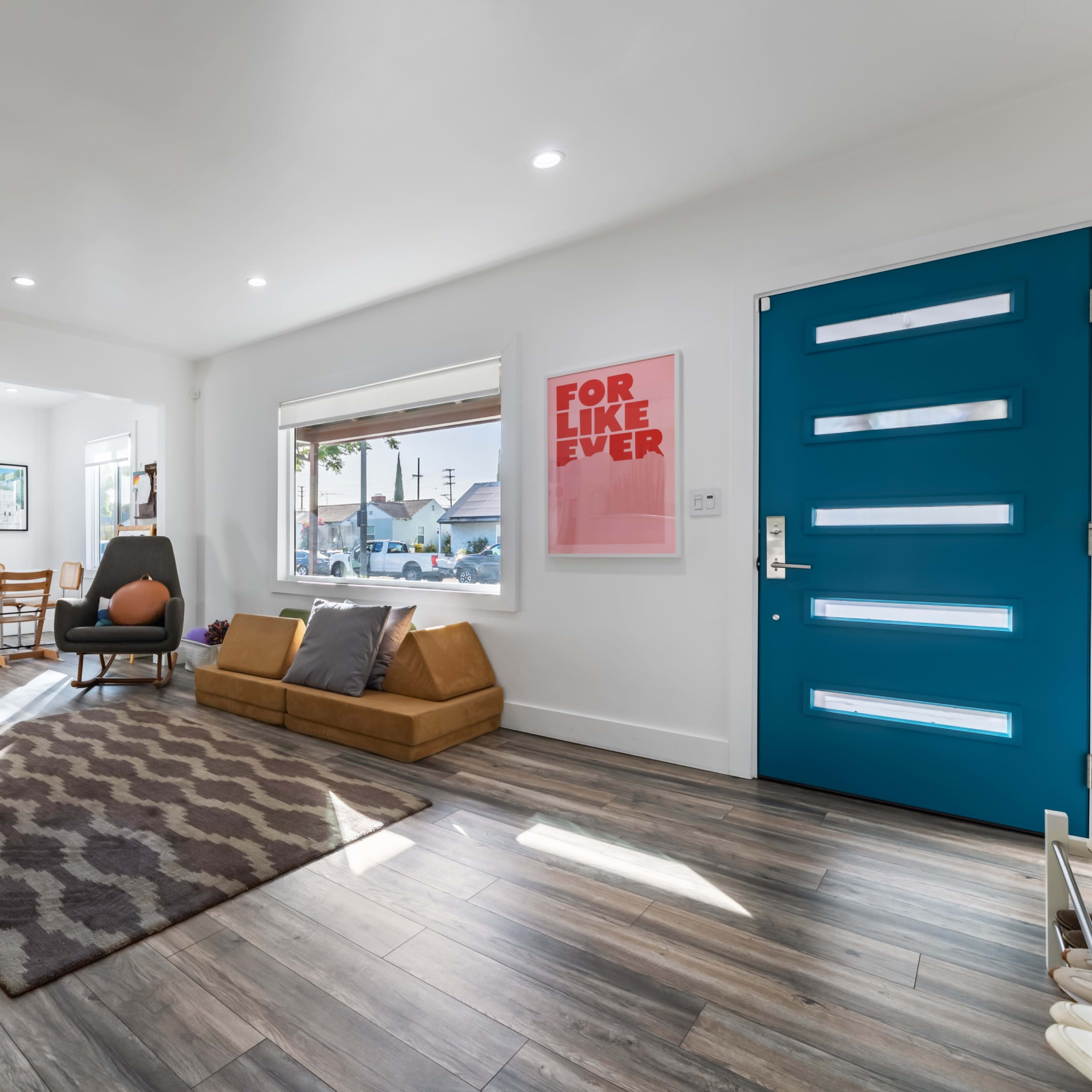 A bright living room features a blue front door, a brown sofa, a patterned rug, and a small dining area with wooden chairs.