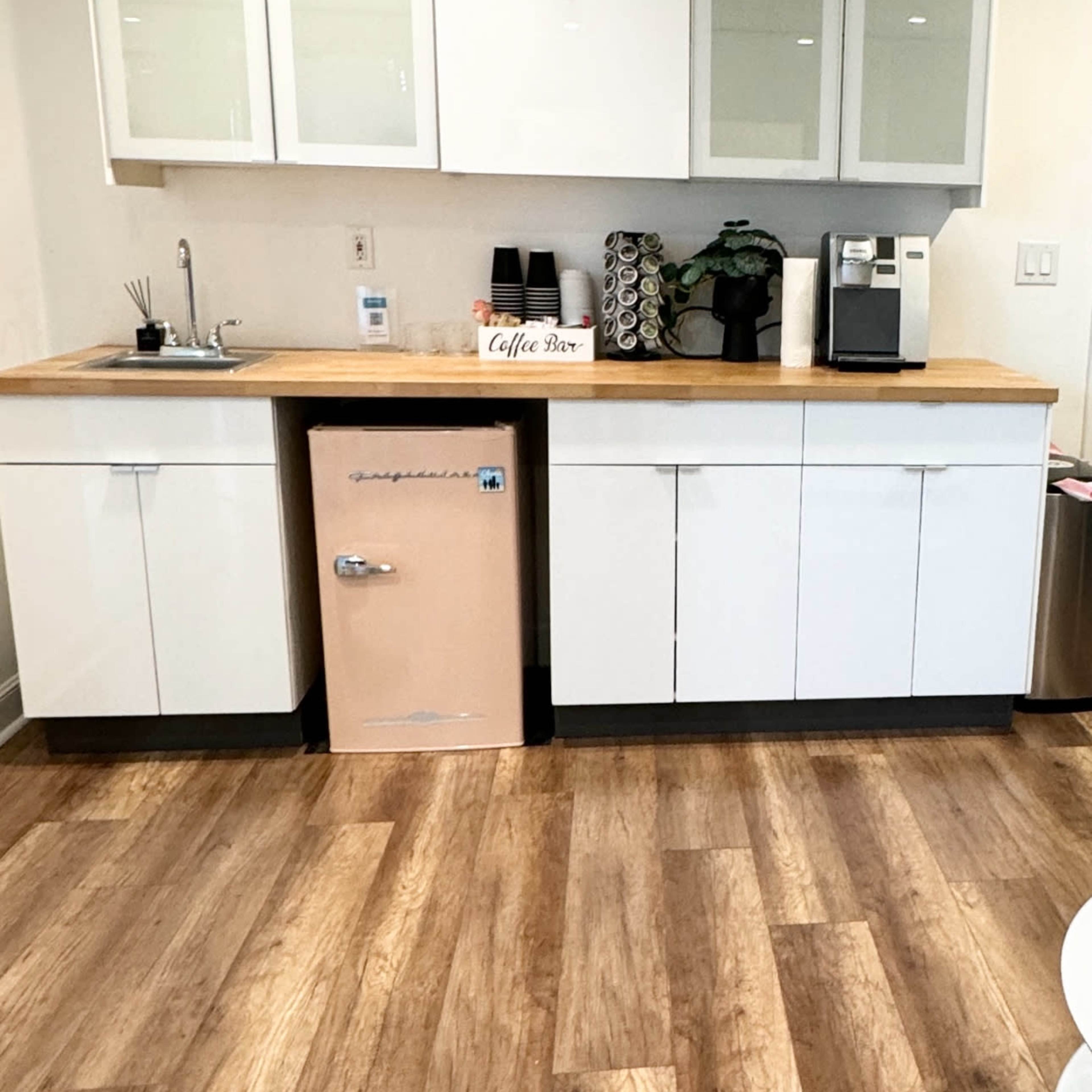A modern kitchen area with white cabinetry, a wooden countertop, a small sink, a pink refrigerator, and a coffee station.
