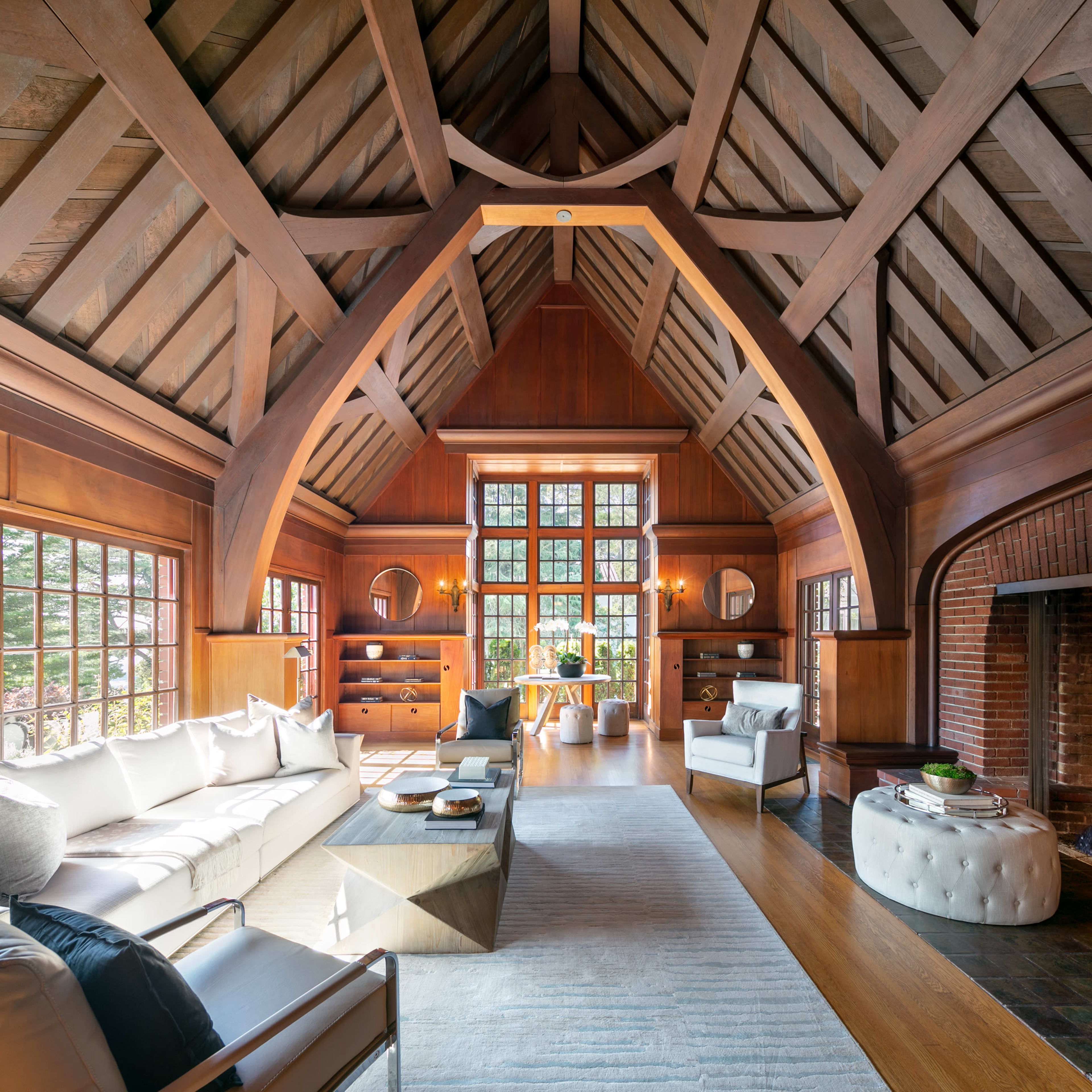 A spacious living room with high wooden-beamed ceilings, large windows, and a fireplace, furnished with modern seating and a coffee table.
