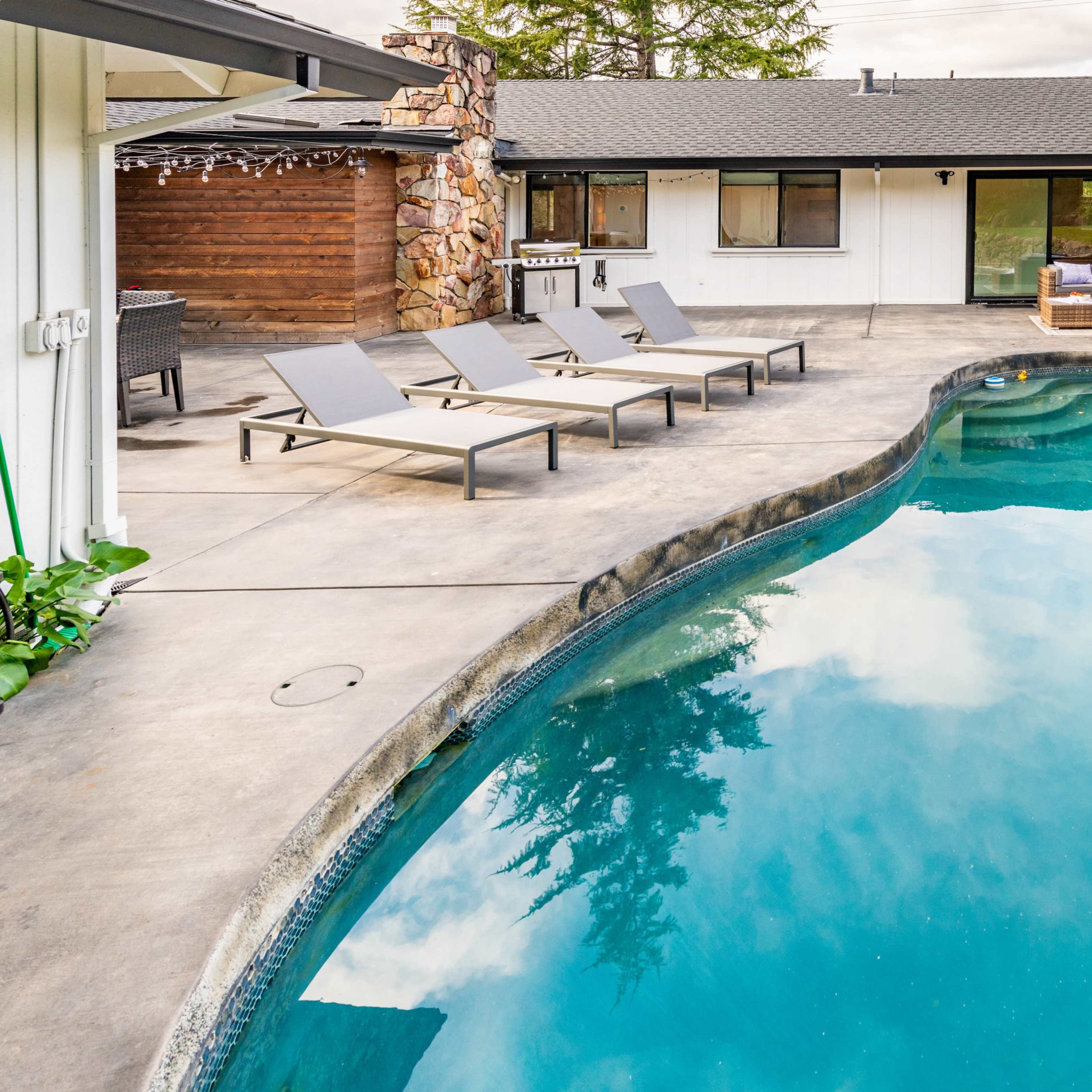 A rectangular swimming pool with lounge chairs surrounds a modern home featuring a stone accent wall and a patio area.